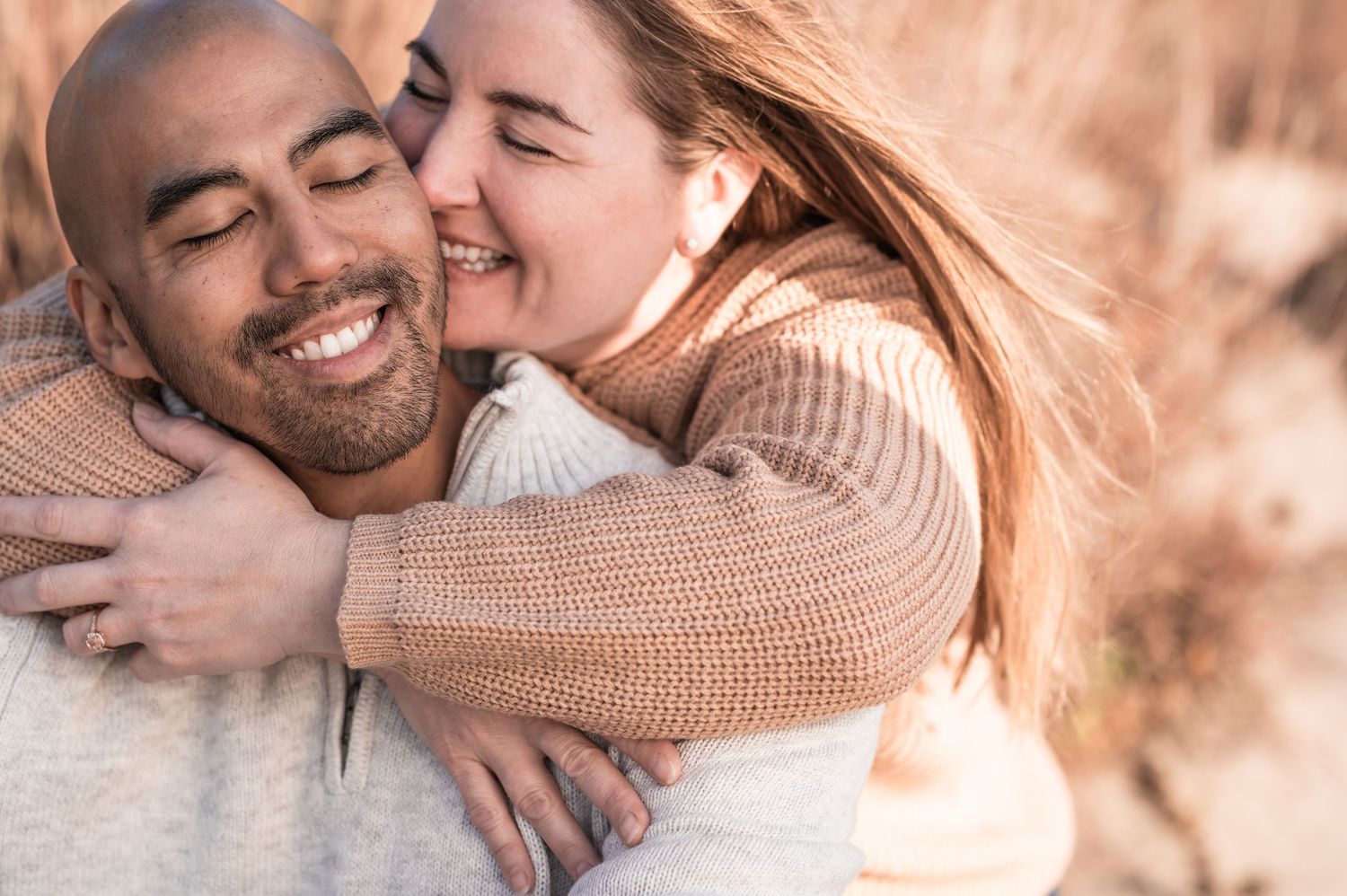 Happy couple embracing outdoors, woman smiling and hugging man from behind in matching beige sweaters.