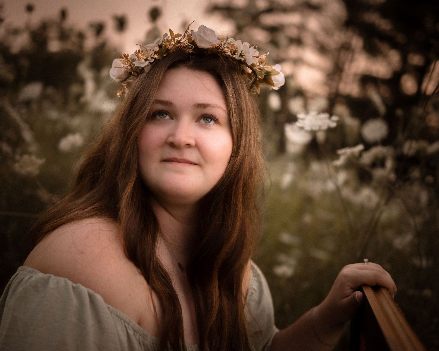 Young woman with floral crown and off-shoulder dress poses in a wildflower field at golden hour.