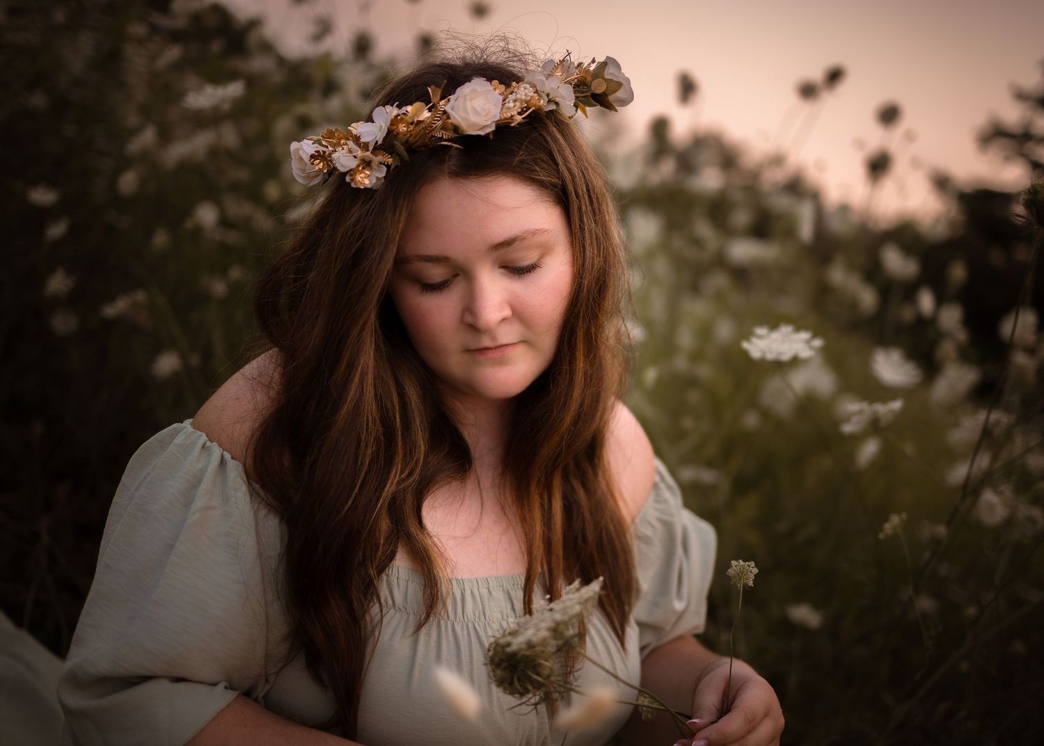Young woman with floral crown holds dandelion in a dreamy wildflower field at golden hour.