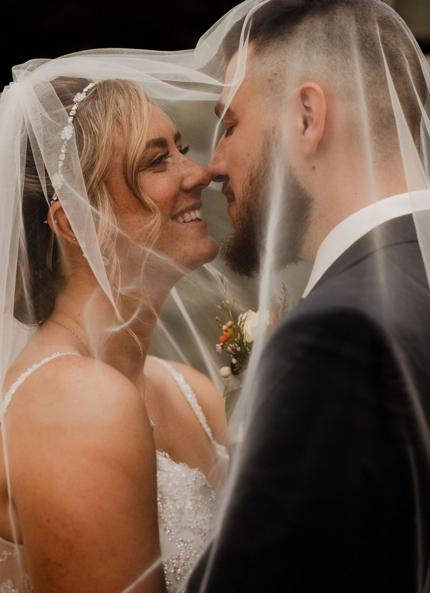 Bride and groom smiling closely together under flowing wedding veil in romantic portrait.