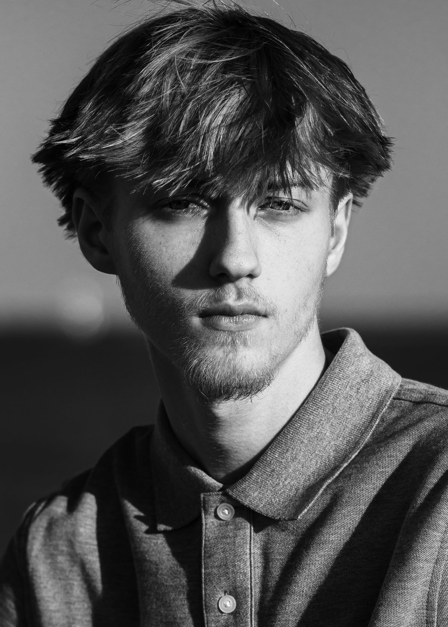 Black and white portrait of a young man with tousled hair and light stubble outdoors.