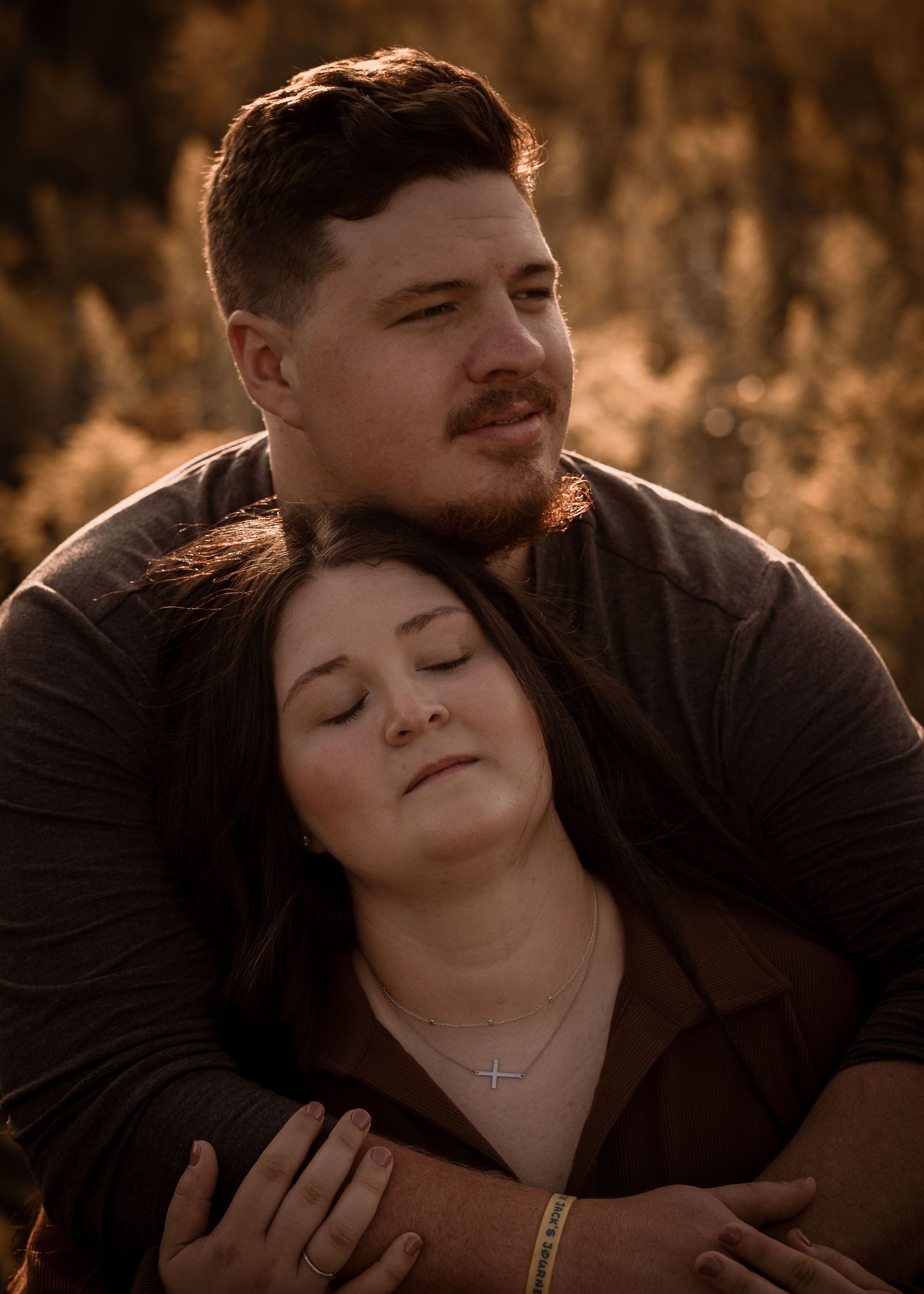 Couple embracing at golden hour, woman wearing cross necklace, man holding her from behind in warm sunset light.