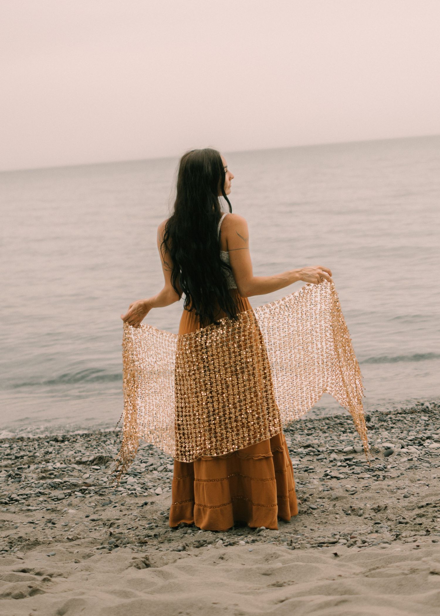 Woman in golden skirt holding lace shawl on rocky beach, long dark hair, facing the ocean.