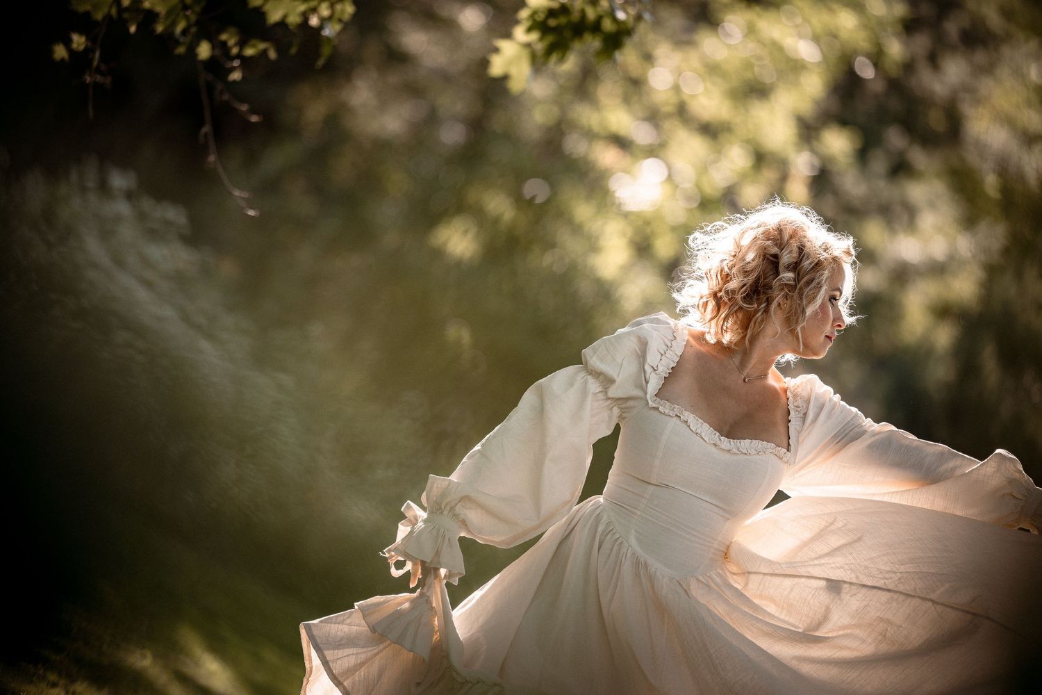 A woman in a flowing white vintage wedding dress twirls gracefully outdoors in soft golden sunlight.