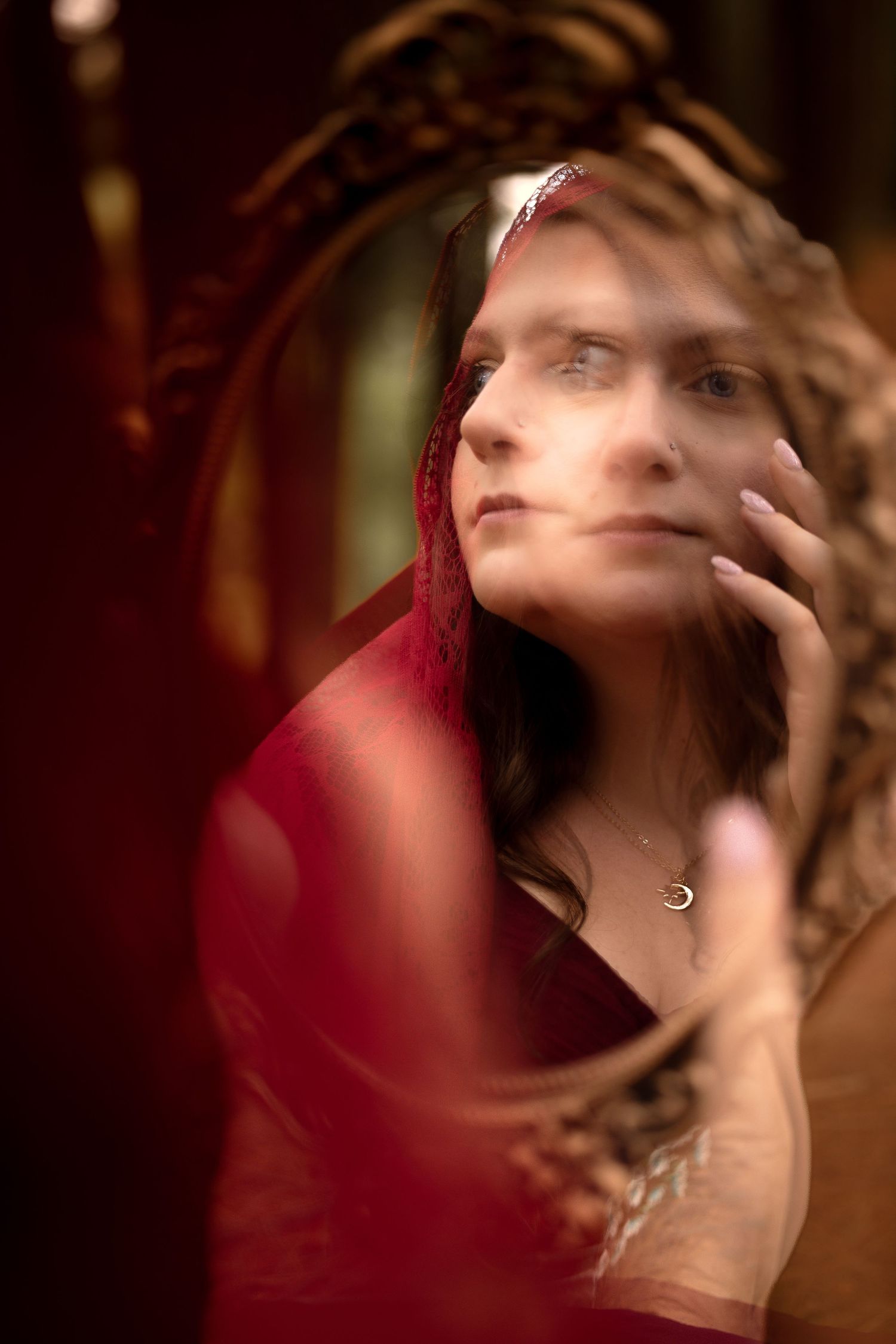 Woman in red sari gazing at ornate mirror reflection, wearing crescent moon necklace and jeweled headpiece.