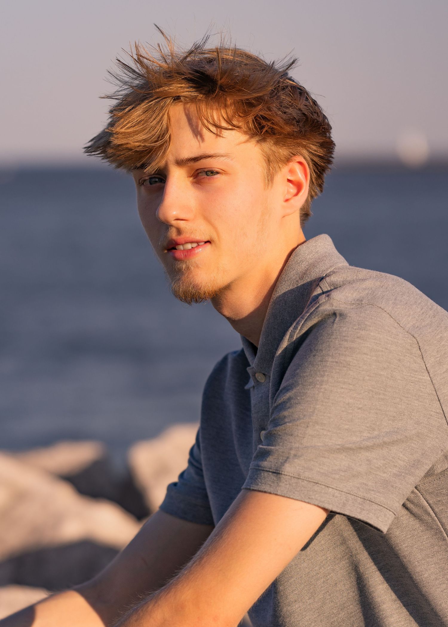 Young man with tousled blonde hair sitting on rocks by the ocean at golden hour sunset.