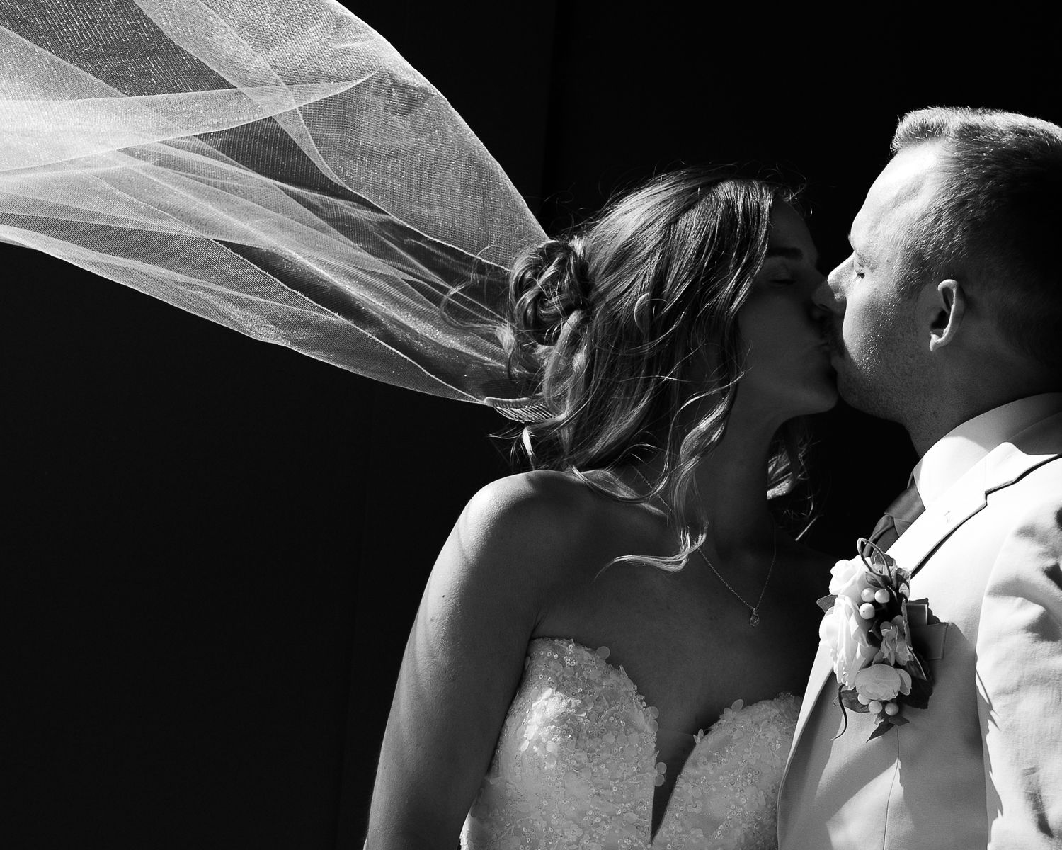 Dramatic black and white photo of couple kissing as bride's veil billows in the wind on wedding day.