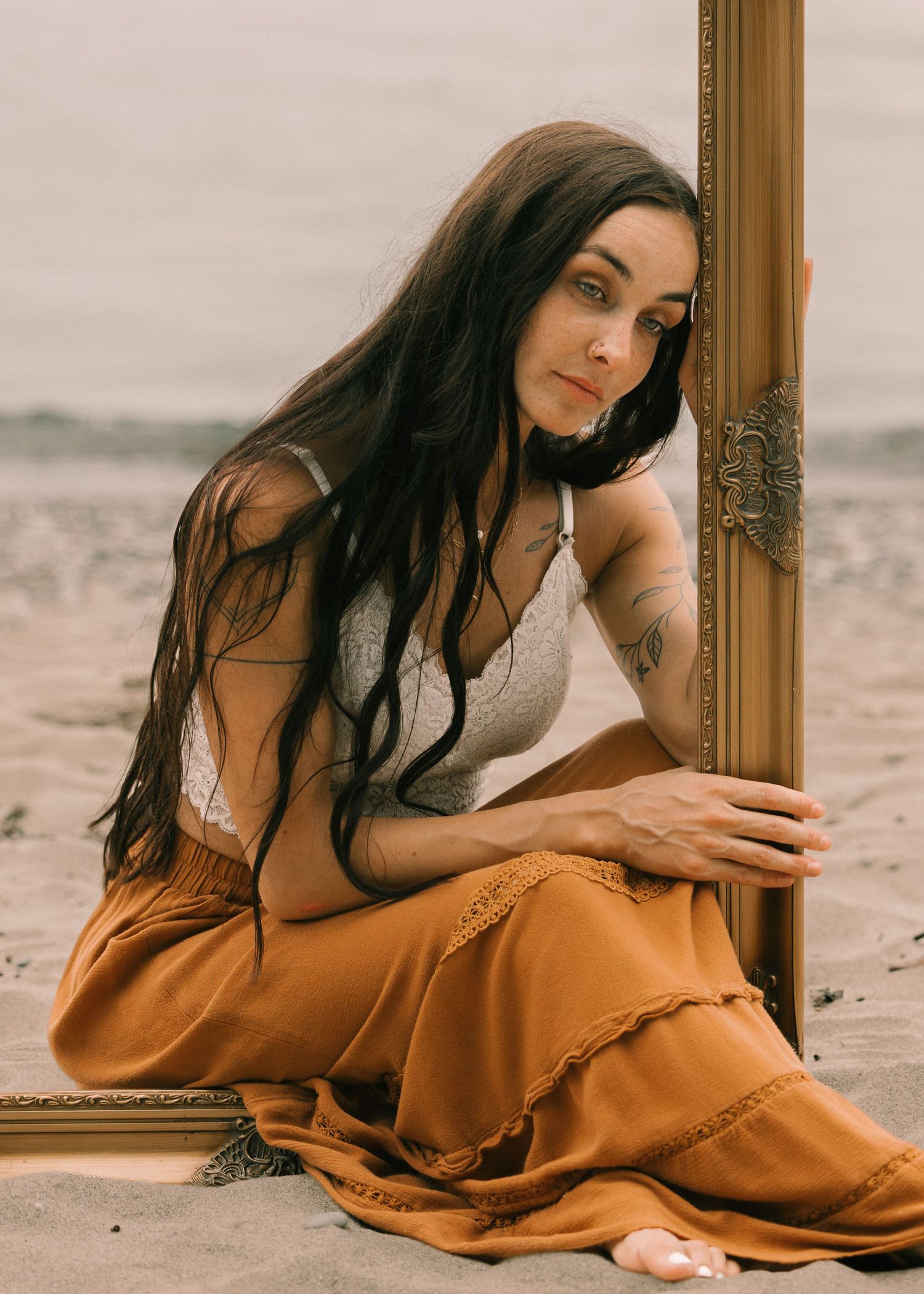 Woman with long dark hair in white top and orange skirt sits on beach leaning against ornate gold picture frame.