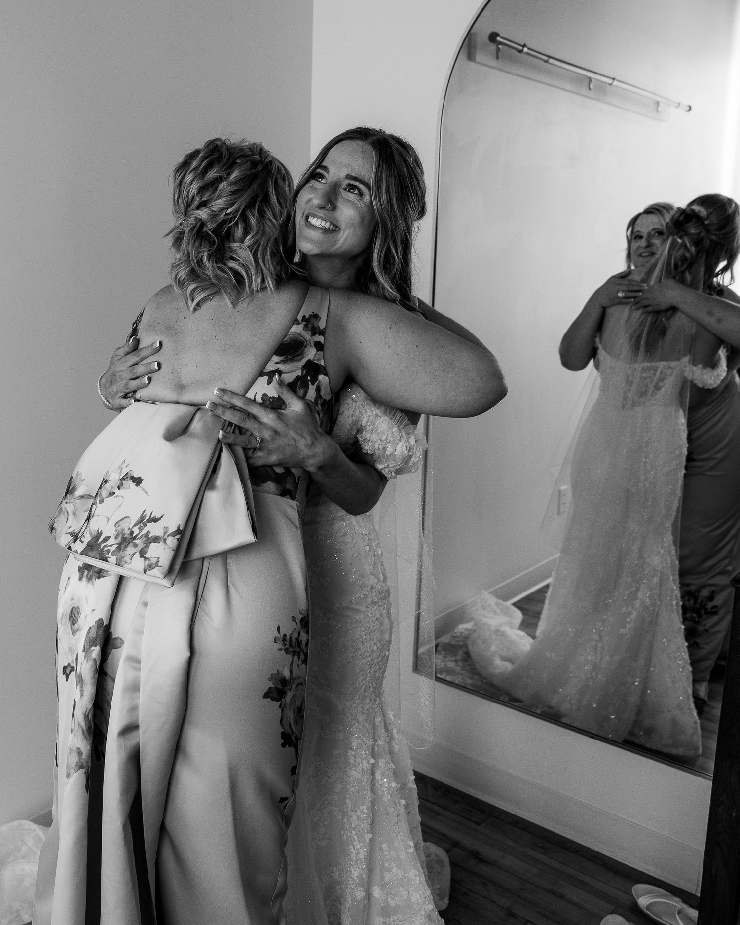 Joyful bride in lace mermaid gown sharing warm embrace with guest, reflected in mirror behind them.