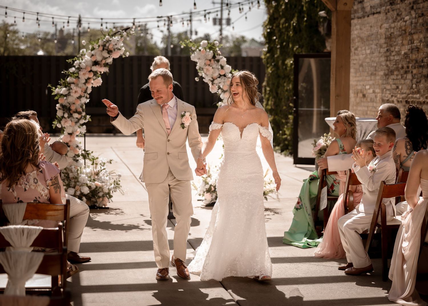 Bride and groom walking down aisle under floral arch at sunny outdoor wedding ceremony.