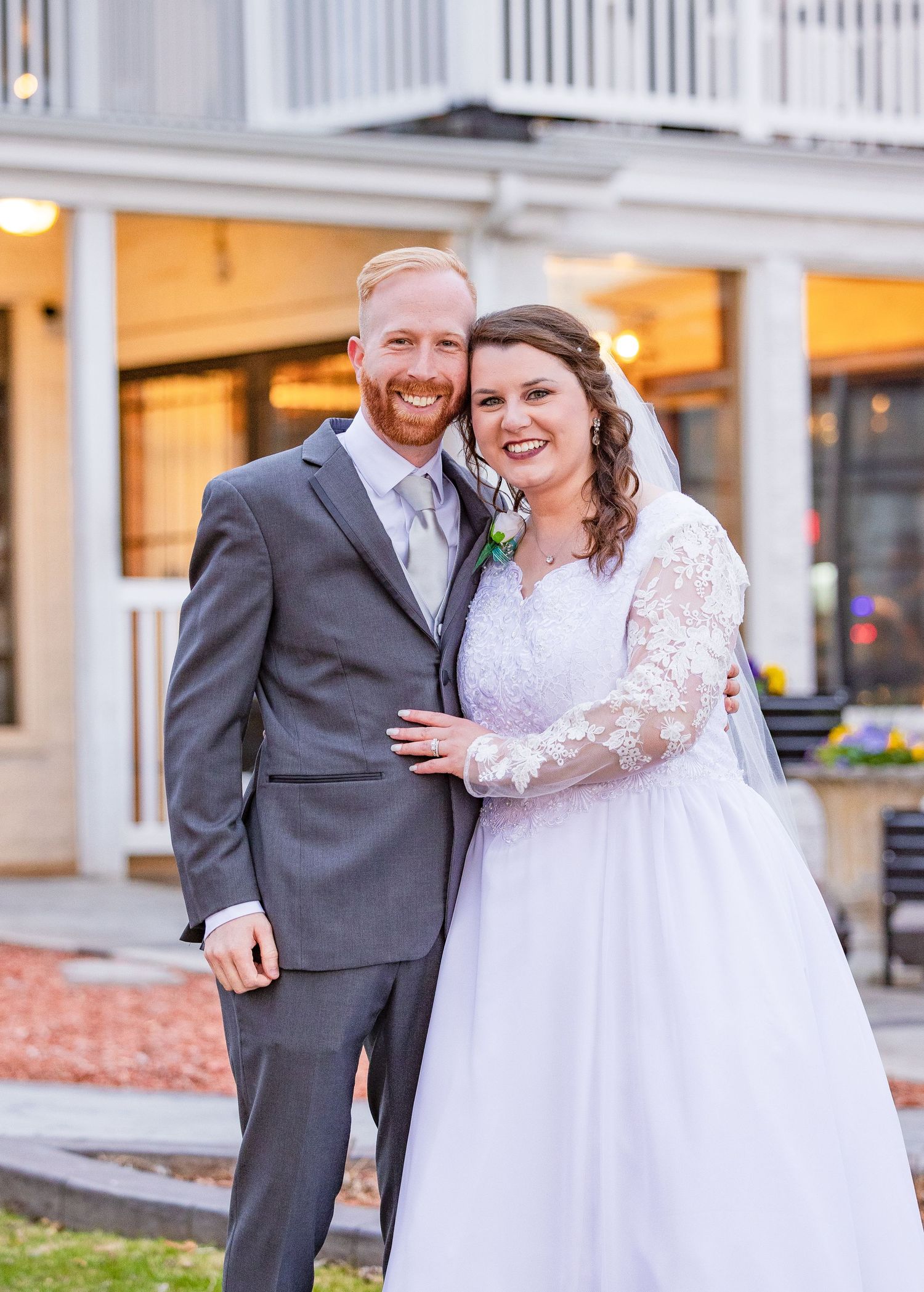 Happy newlywed couple poses outdoors, groom in gray suit and bride in lace wedding gown.