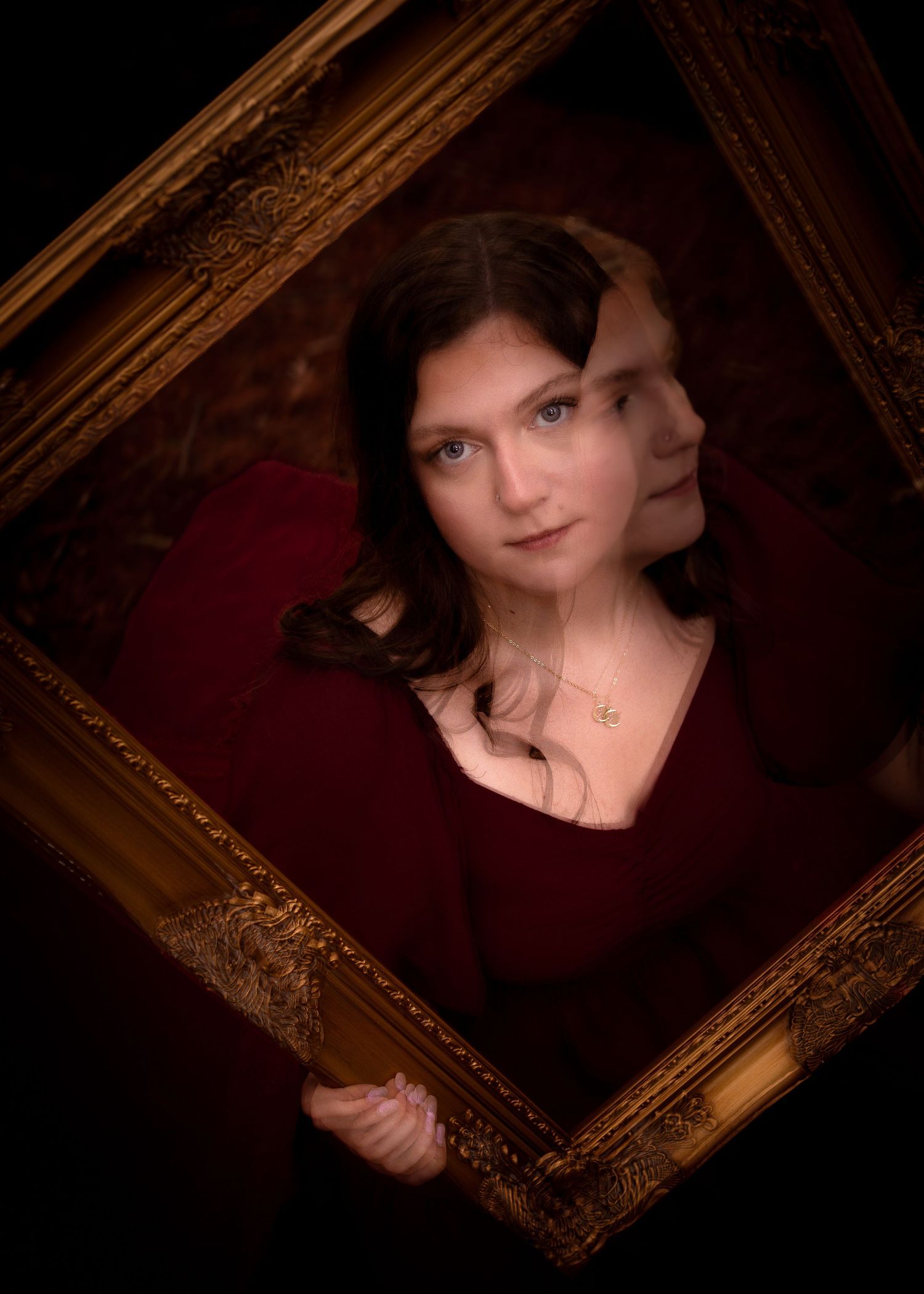 Woman in red dress holding ornate gold picture frame with her reflection visible, dramatic dark background.