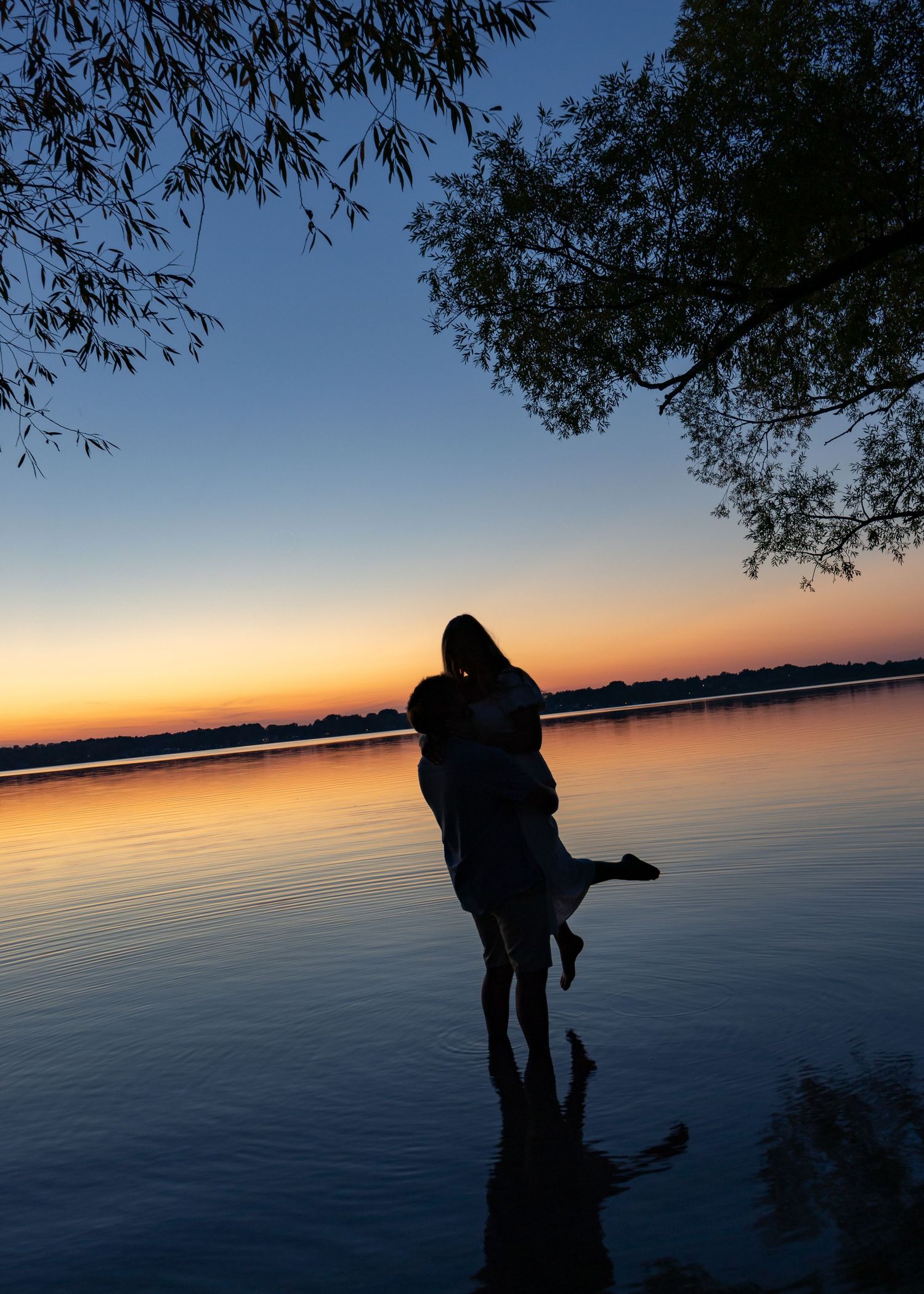 Romantic couple silhouette embracing at sunset by a calm lake with vibrant orange and blue sky reflections.