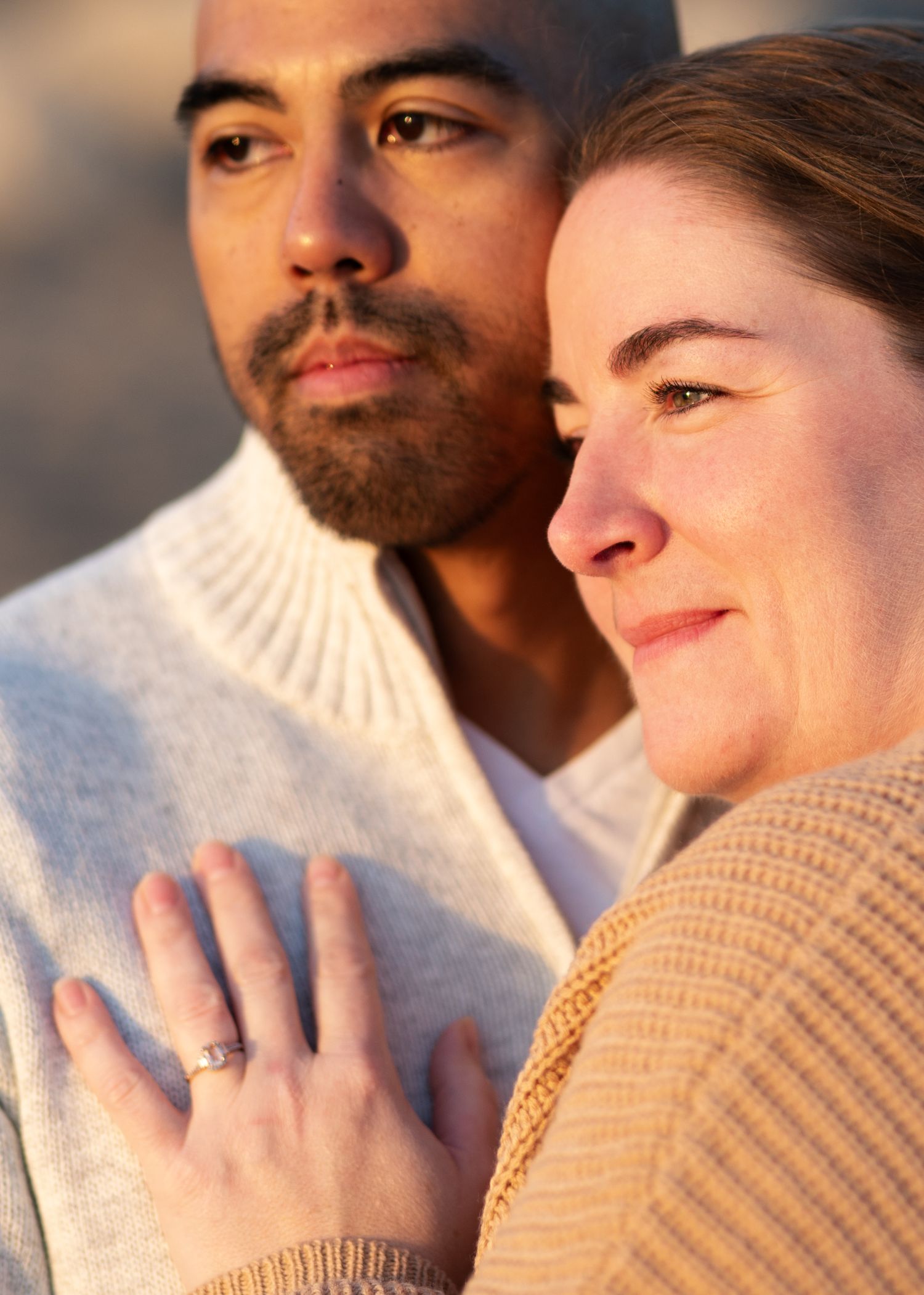Interracial couple posing closely together in warm golden light, woman displaying engagement ring on hand.