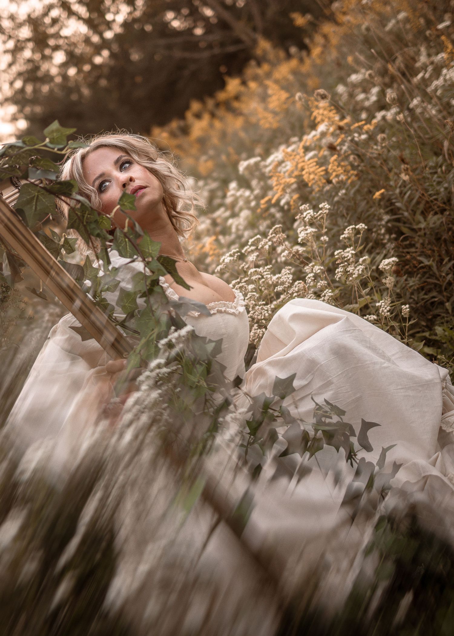 A blonde woman in a white gown sits among wildflowers and ivy in a dreamy, softly lit outdoor setting.