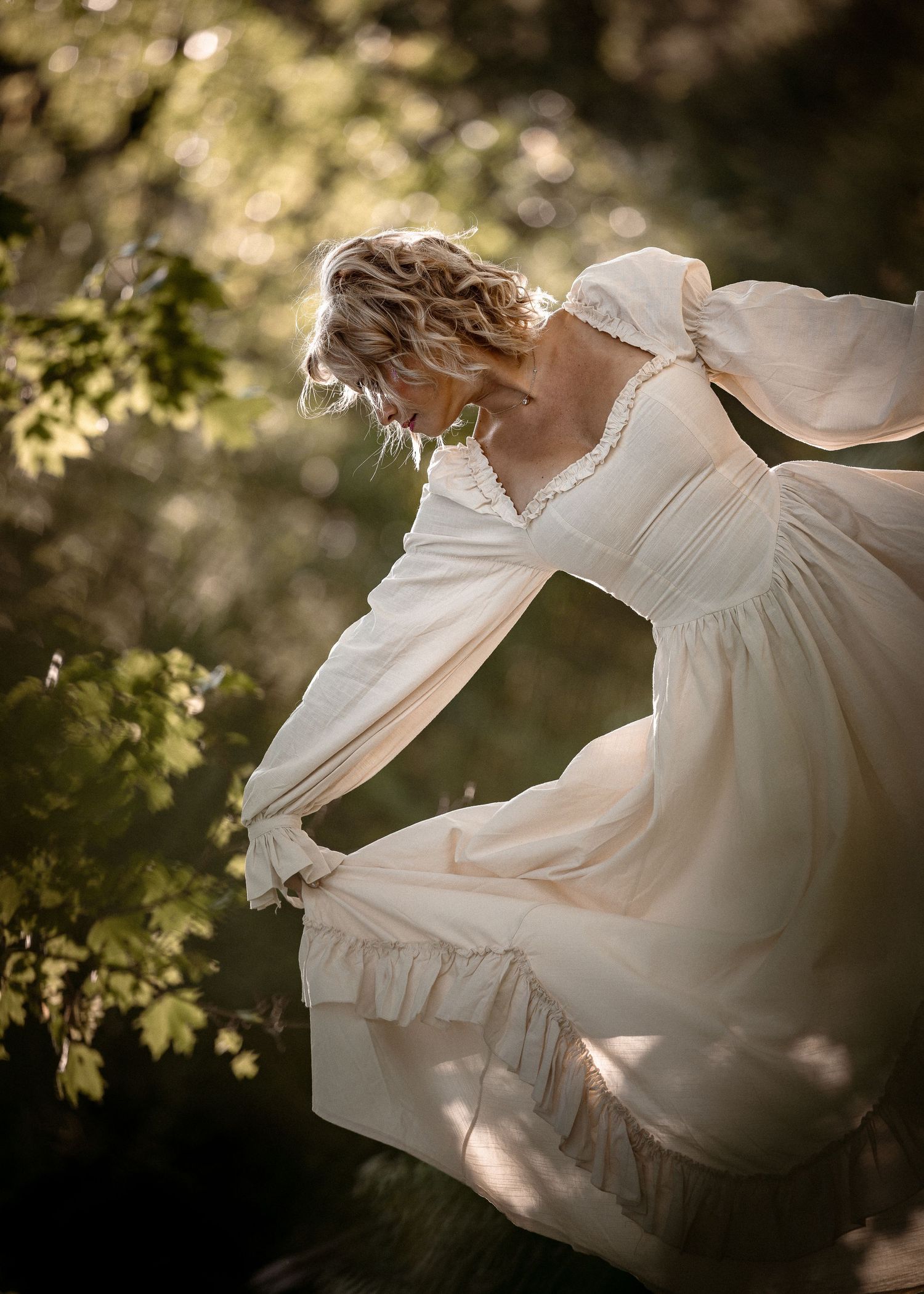 Woman in flowing cream vintage gown twirling gracefully among sunlit green trees in an enchanted forest setting.