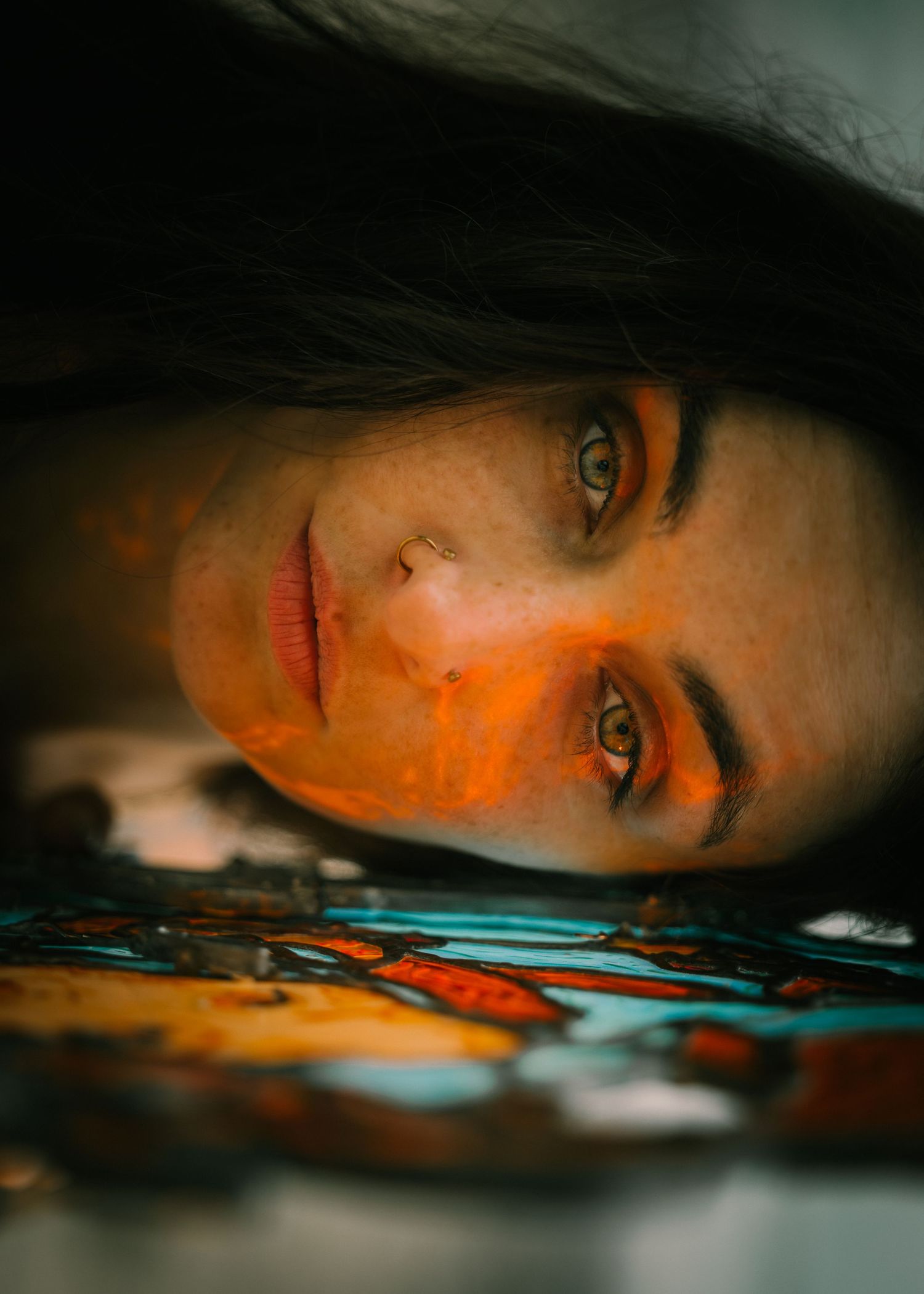 Close-up portrait of a woman with orange paint on her face, lying near colorful stained glass artwork.