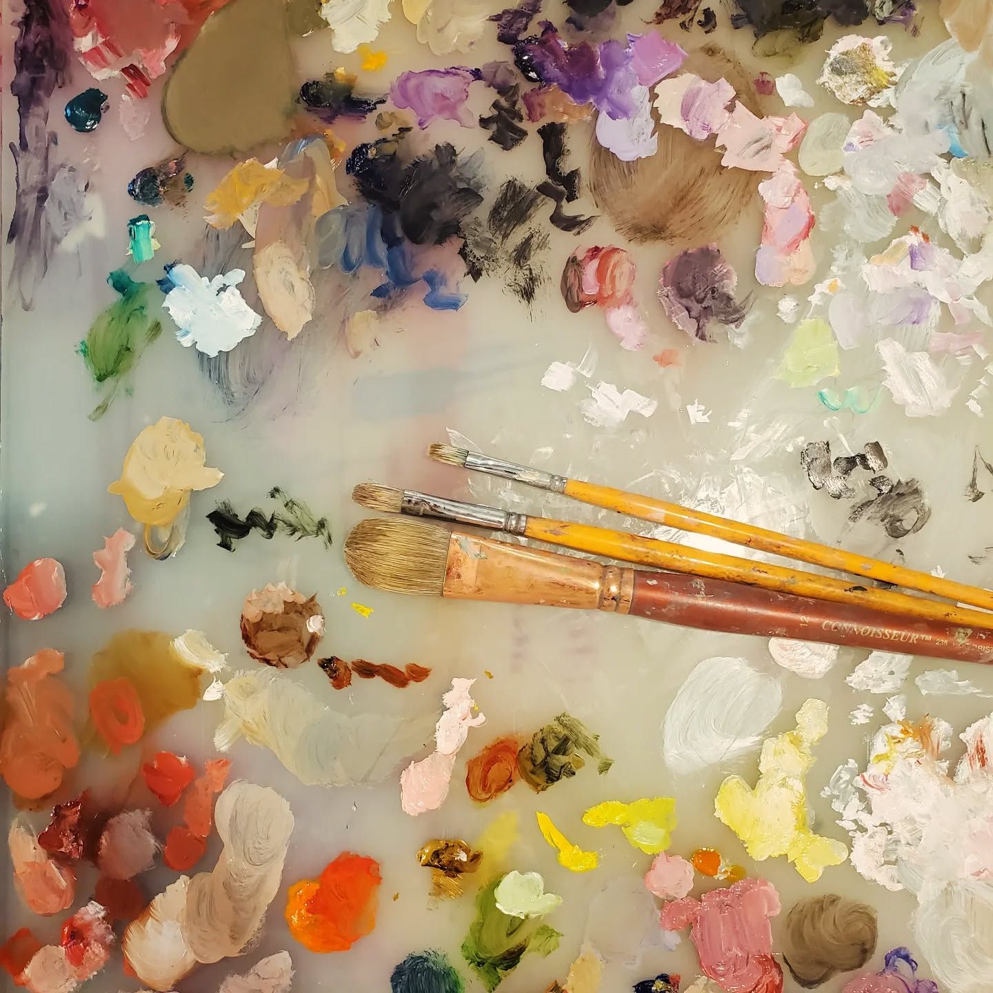 Artist paint palette with multiple brushes resting on surface covered in colorful mixed oil paint swatches.