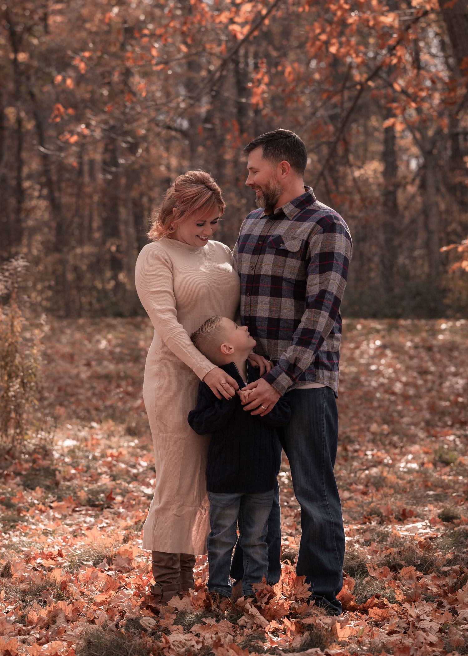 Family of three embraces in autumn forest with colorful fallen leaves on the ground.