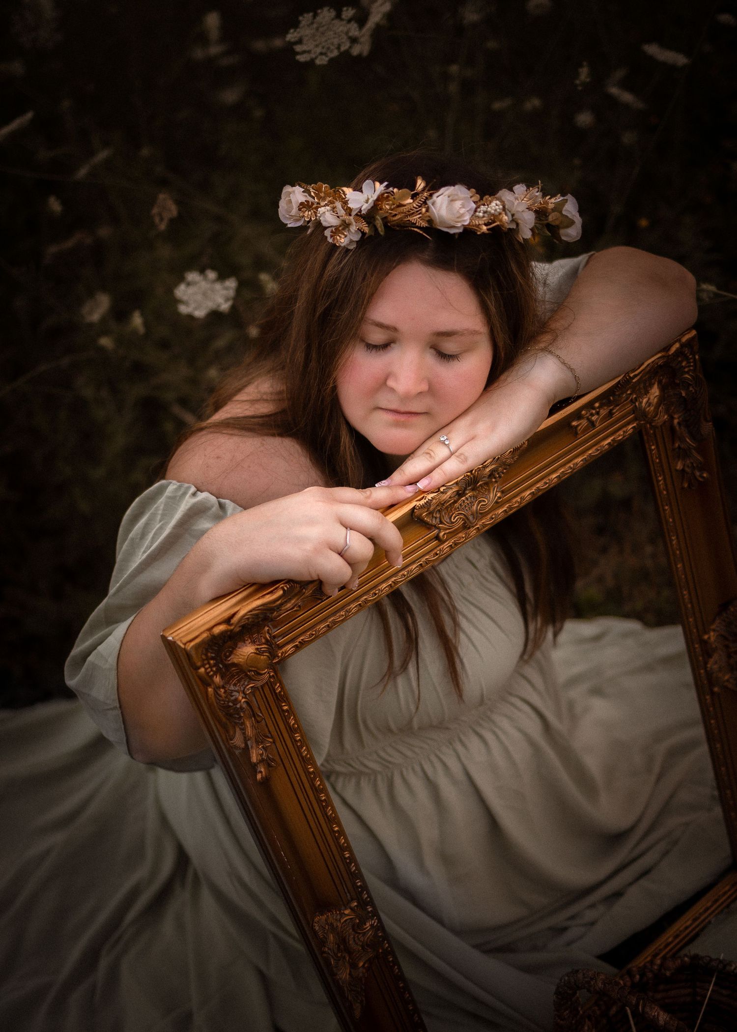 Woman in sage dress and floral crown leans on ornate gold frame outdoors among white blooms.
