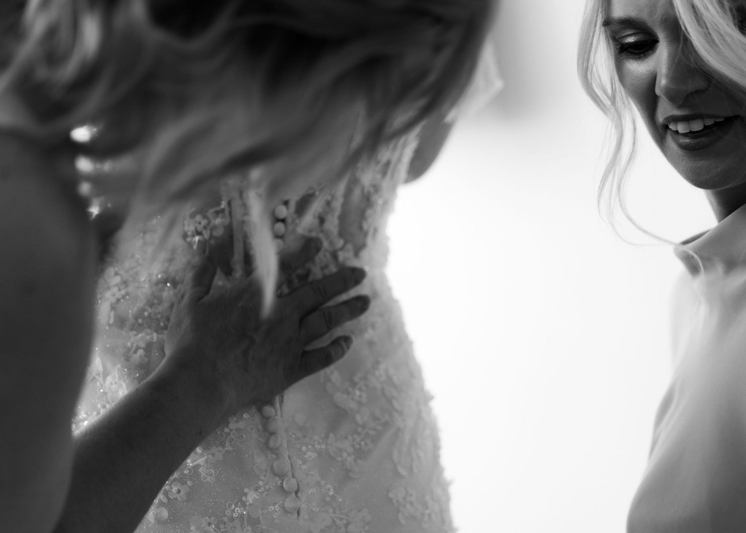 Black and white photo of bride in lace wedding gown being helped by bridesmaid during wedding preparations.