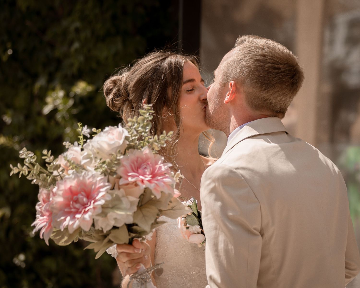 Bride and groom sharing tender kiss while bride holds pink dahlia bouquet outdoors.