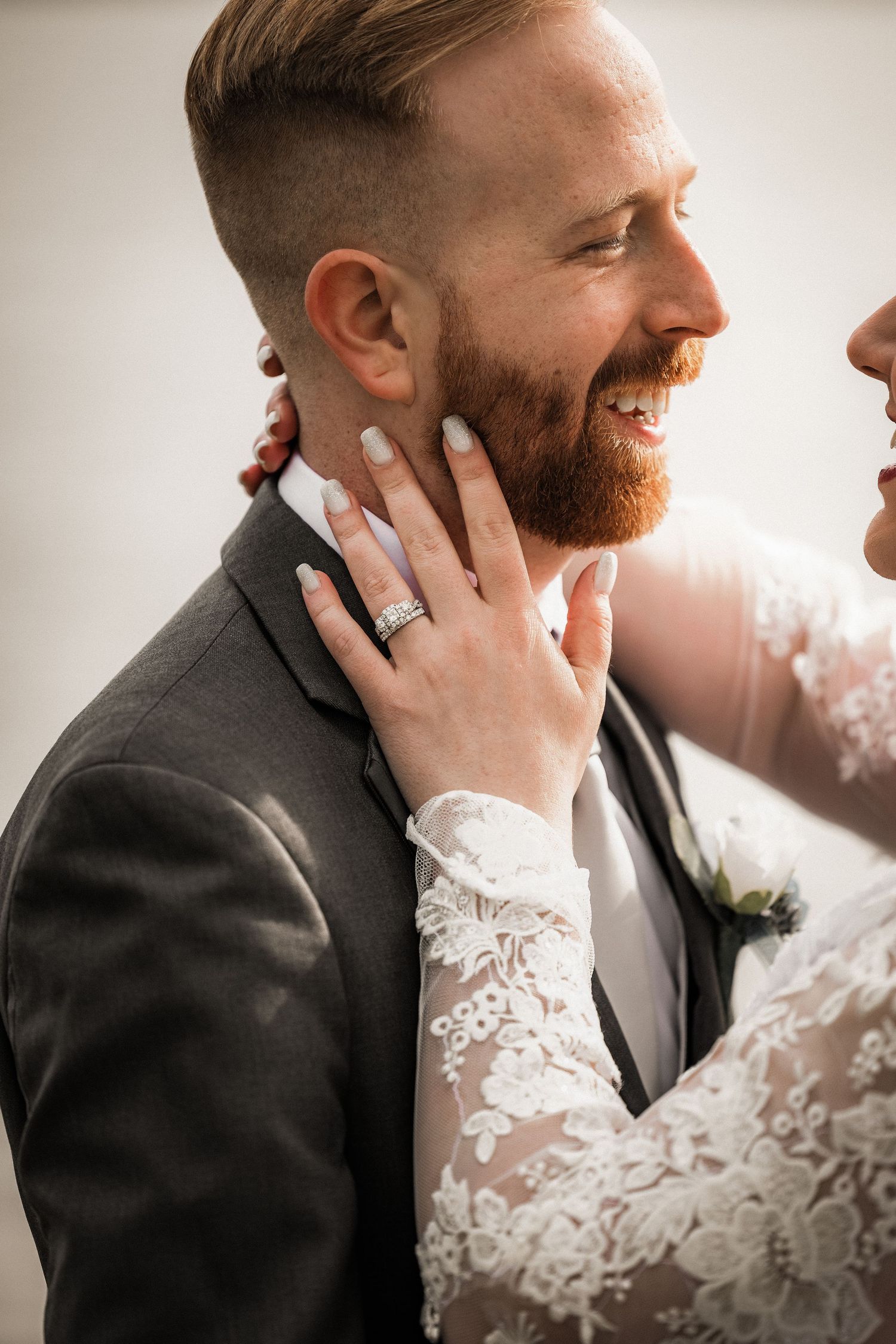 Smiling groom with red beard in gray suit as bride in lace dress tenderly touches his face on wedding day.
