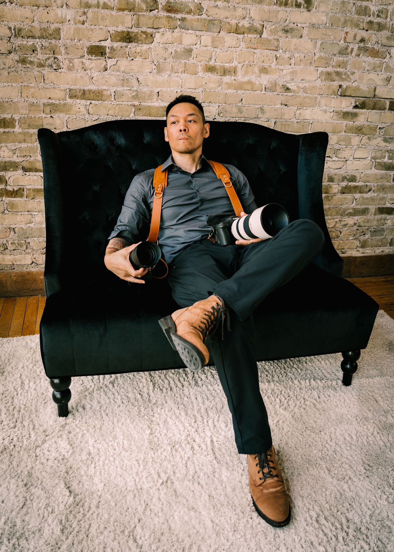 Photographer sitting relaxed in a velvet chair holding cameras with leather harness against brick wall.