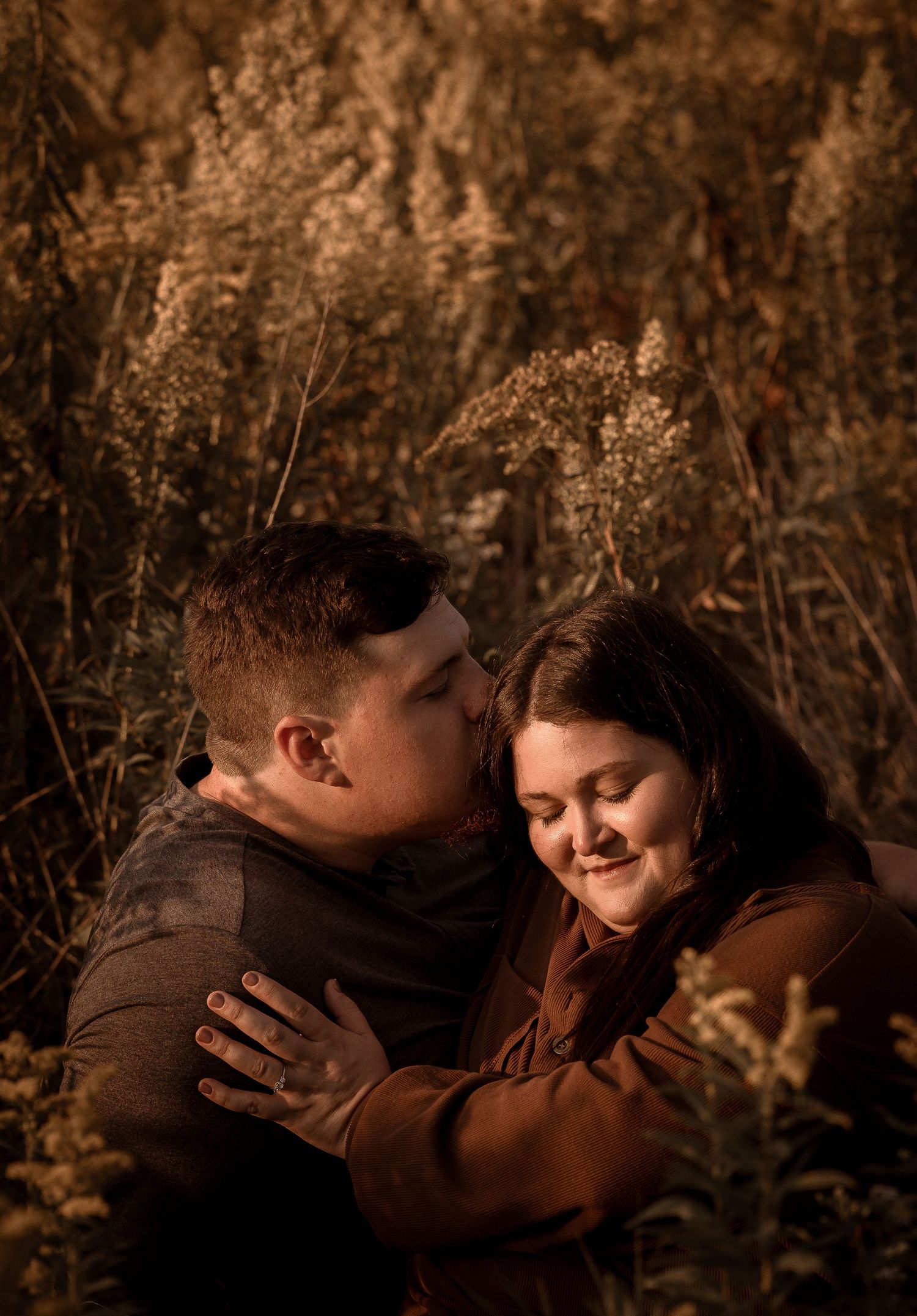 Couple embracing in golden autumn field, man nuzzling woman's cheek as she smiles softly among tall dry grasses.