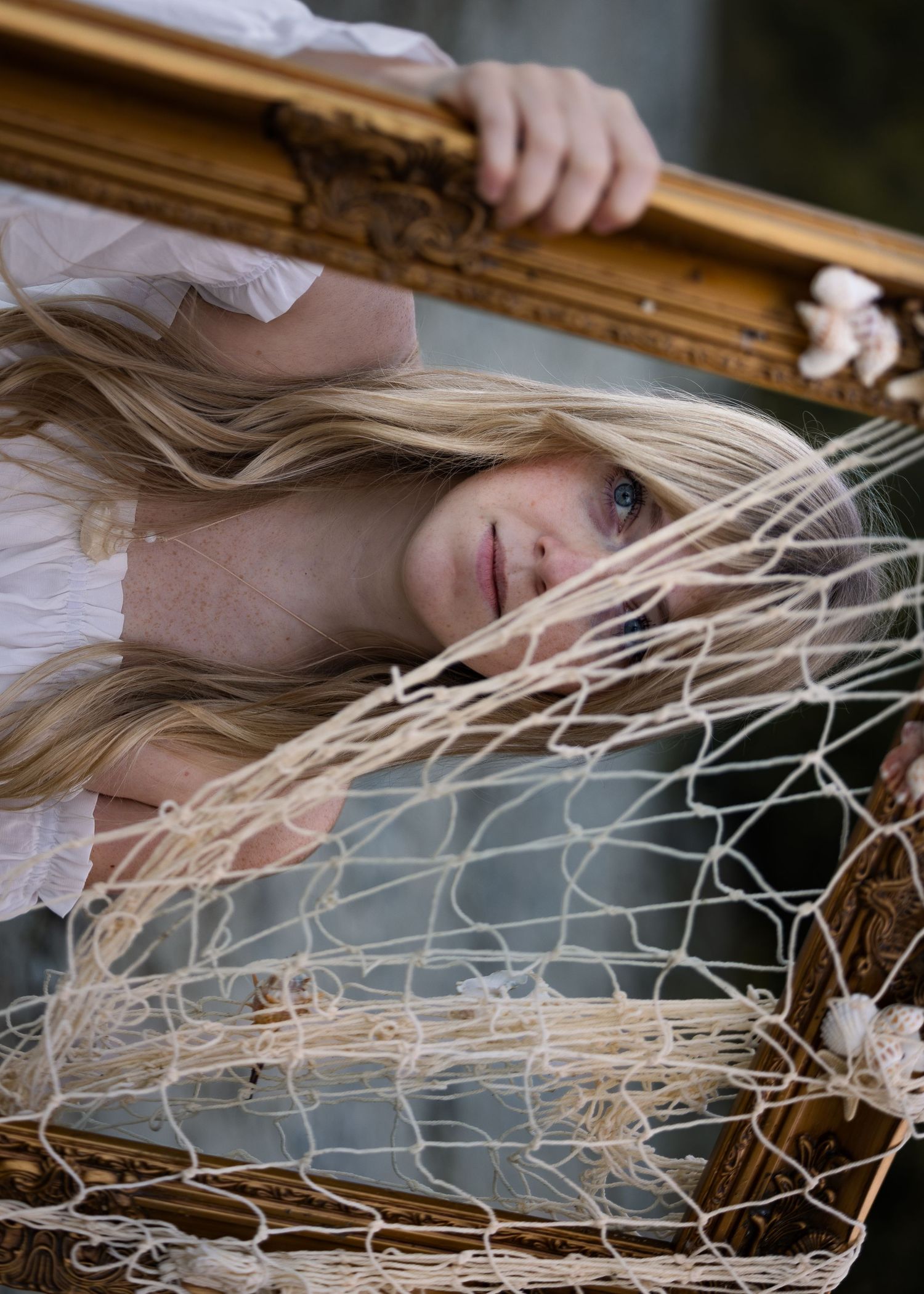 Blonde woman lying amid a woven net inside a gilded frame, wearing a white dress in an artistic portrait.