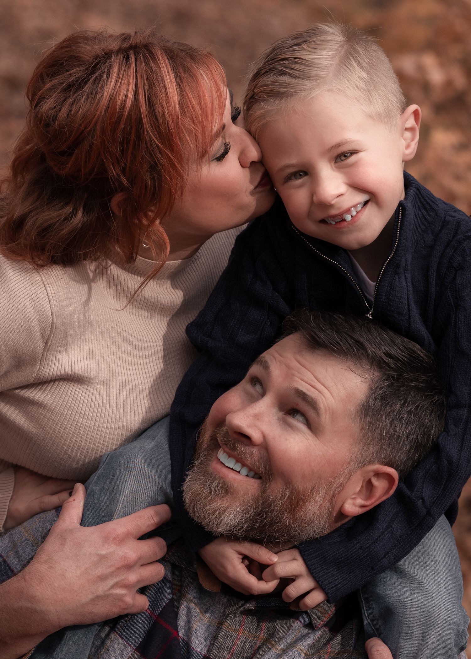 Mother kisses young son while father smiles below in a cozy outdoor family portrait.