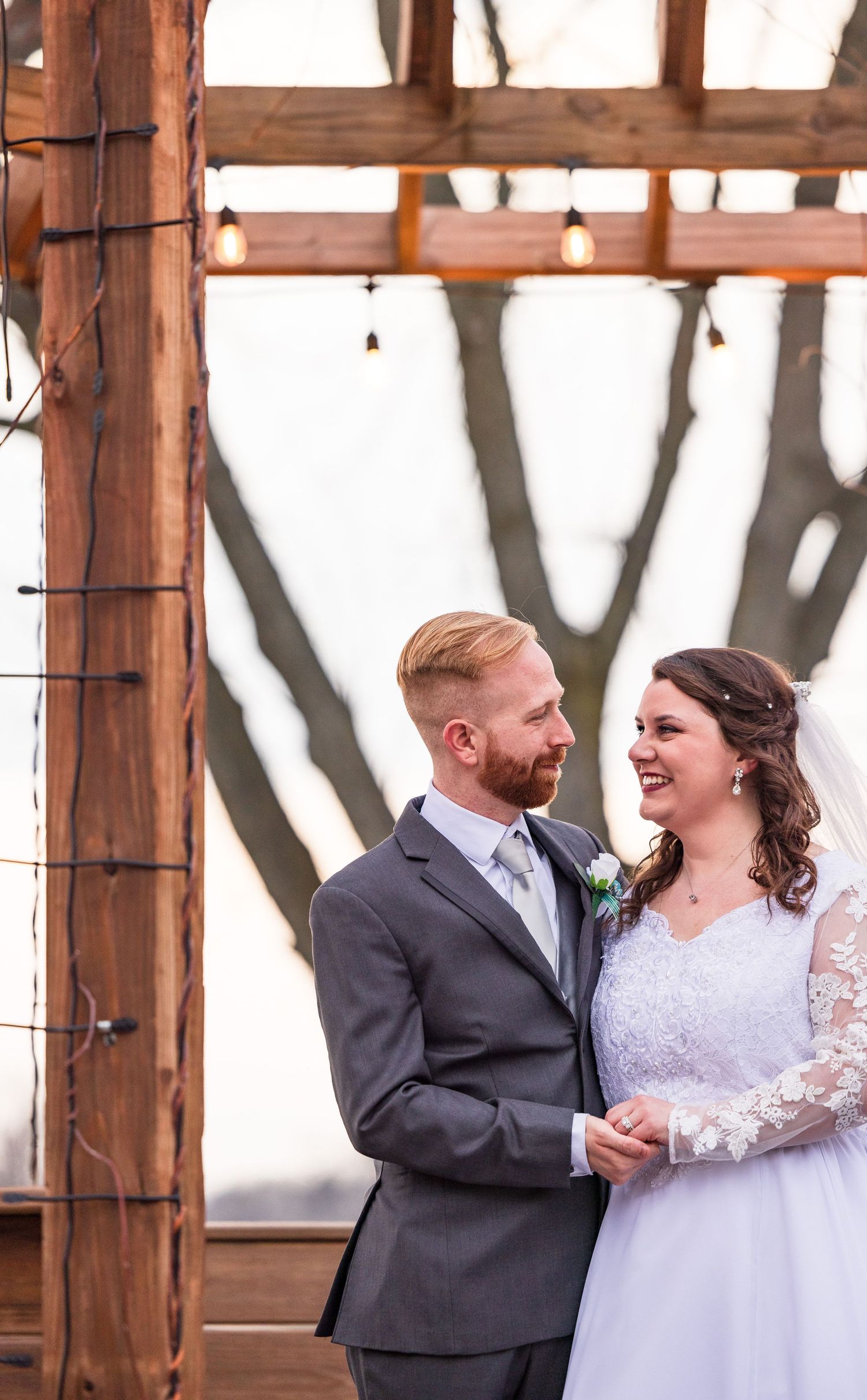 Bride in white gown and groom in gray suit embracing by a serene lakeside on their wedding day.