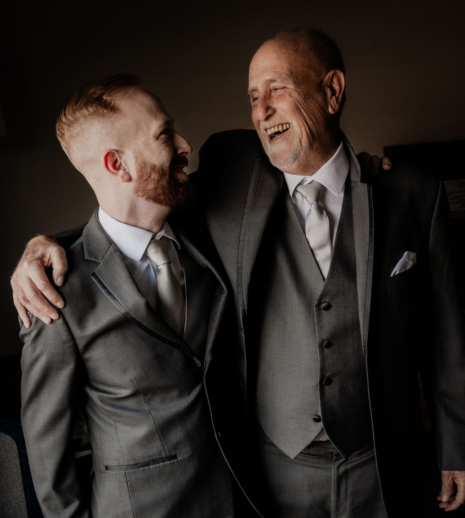 Groom and father sharing warm laugh together in matching gray suits before wedding ceremony.