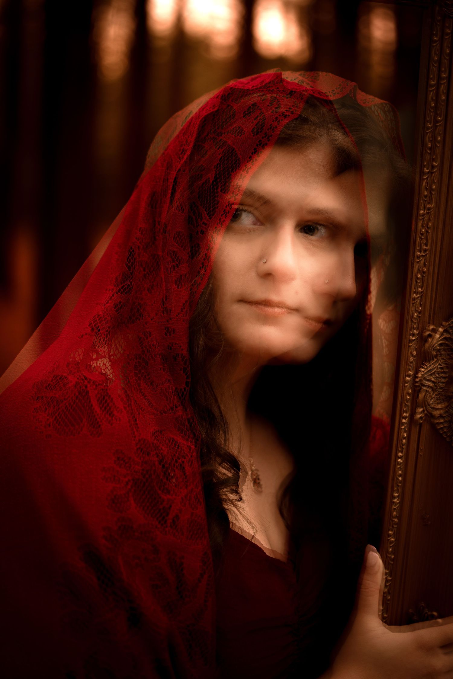 A mysterious woman draped in a red lace veil gazes upward while holding an ornate golden mirror frame.