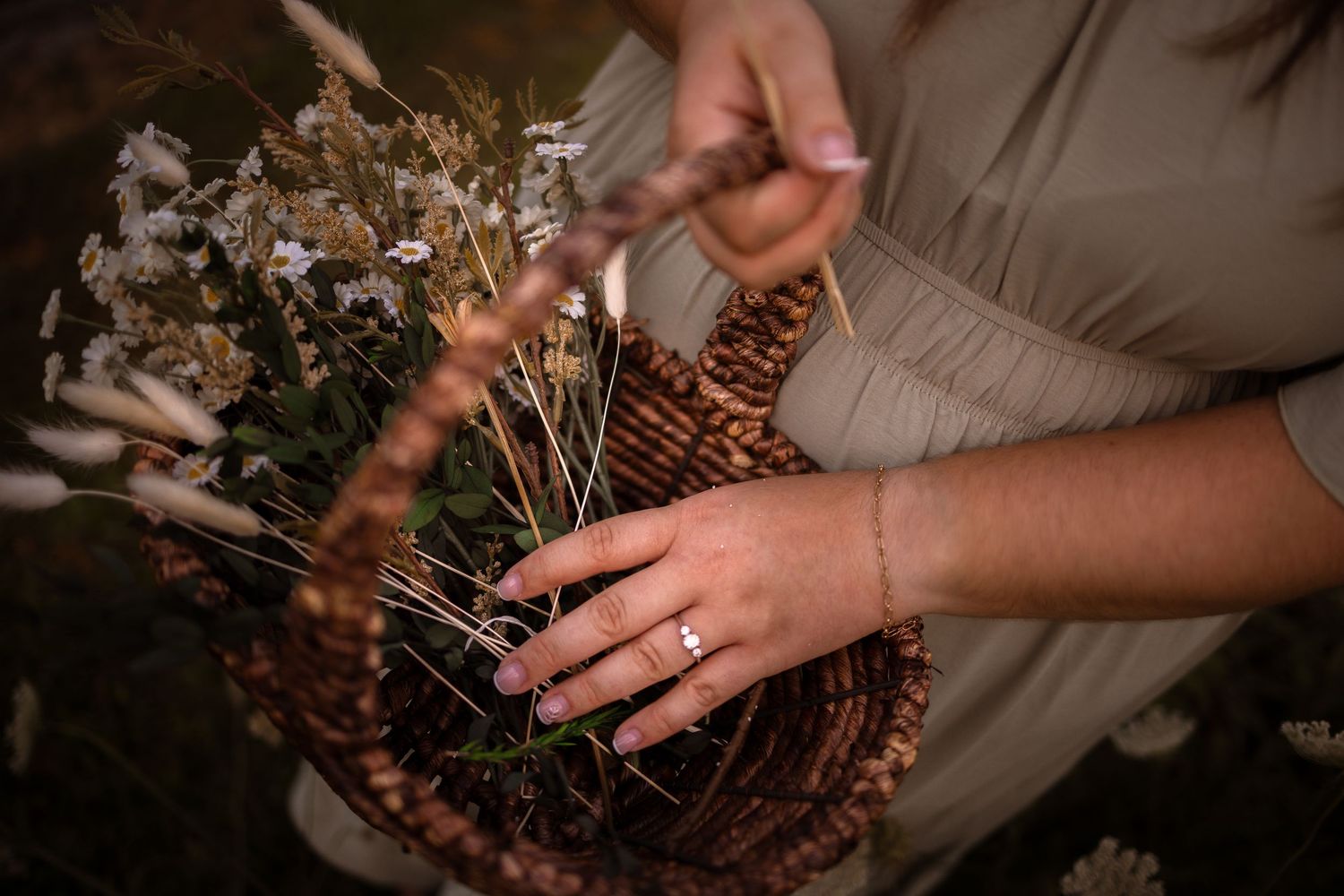 Woman arranging dried wildflowers in a woven basket, wearing a delicate ring and bracelet.