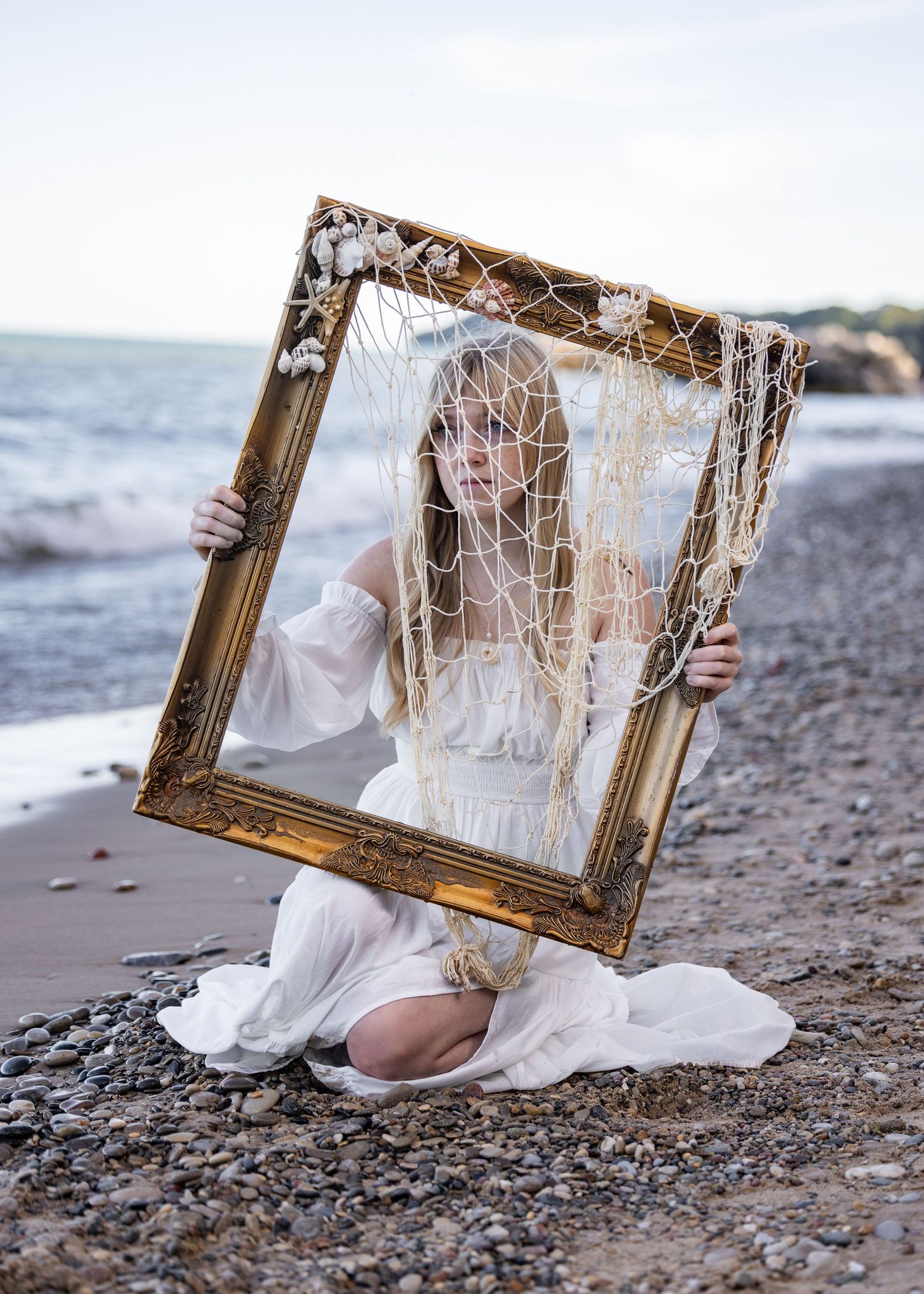 A smiling woman in a white dress holds a gold picture frame with netting on a rocky beach by the ocean.