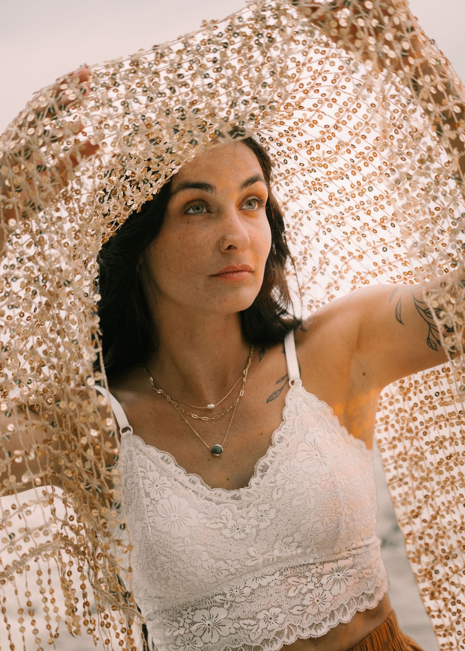Woman in white lace crop top draped in gold sequin fabric, wearing layered necklaces and tattoo, gazing upward outdoors.