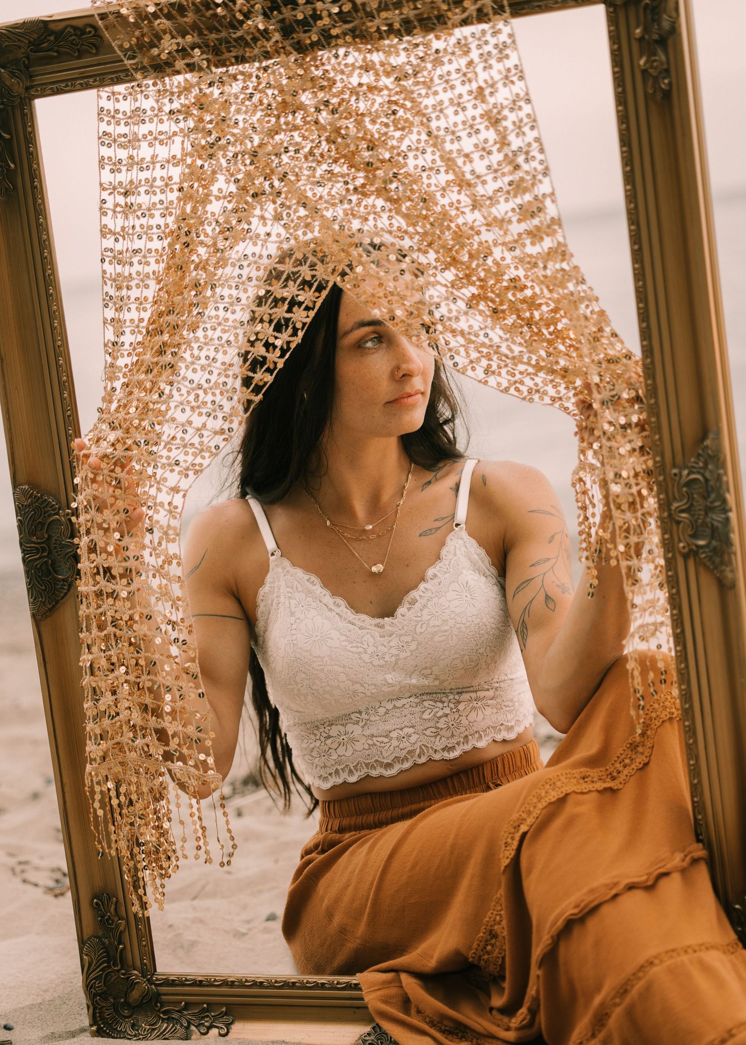 Woman in white lace top and gold skirt poses inside ornate frame with golden beaded veil outdoors.