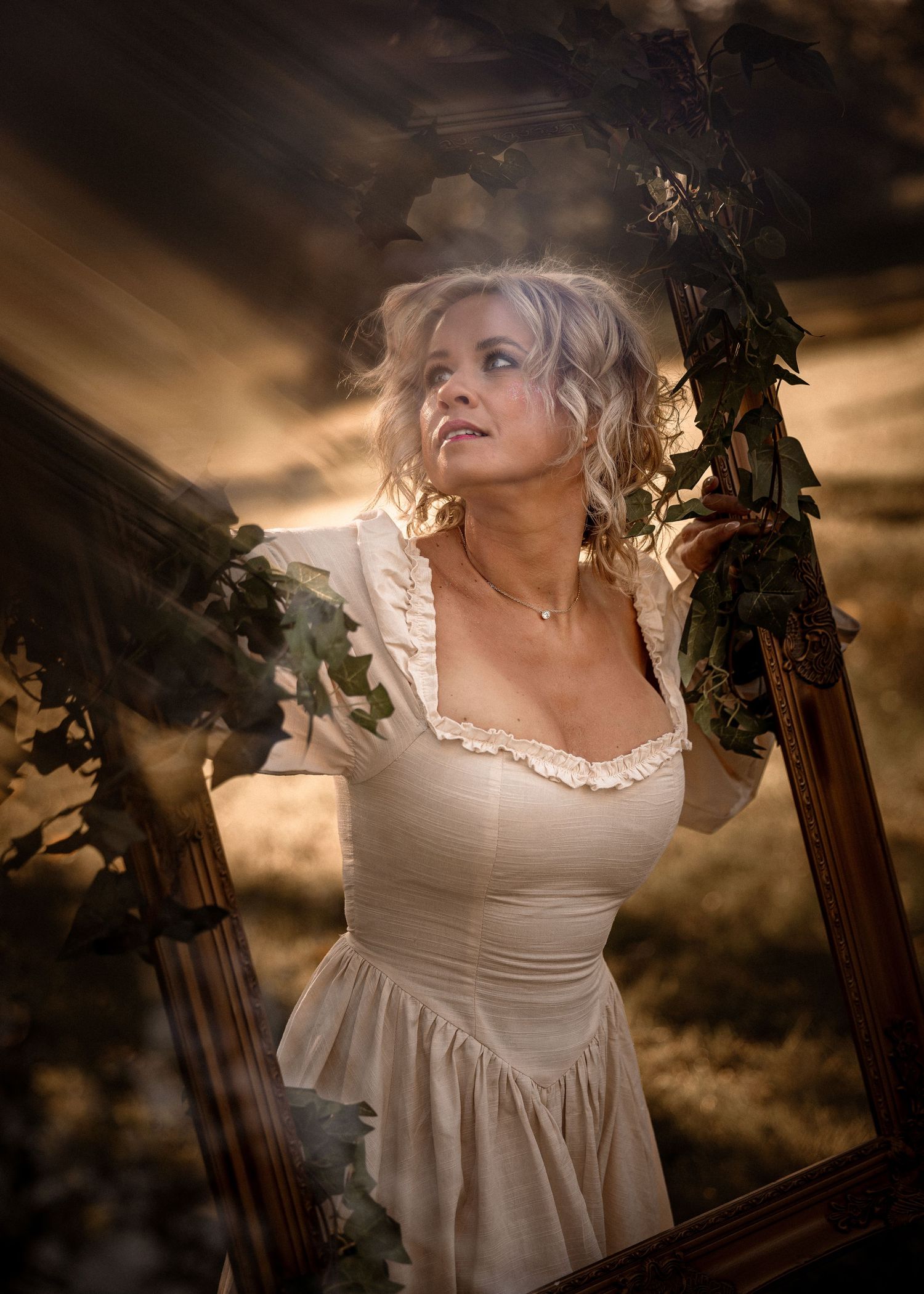 Blonde woman in vintage cream dress holding an ornate frame with ivy, dramatic golden sky background.