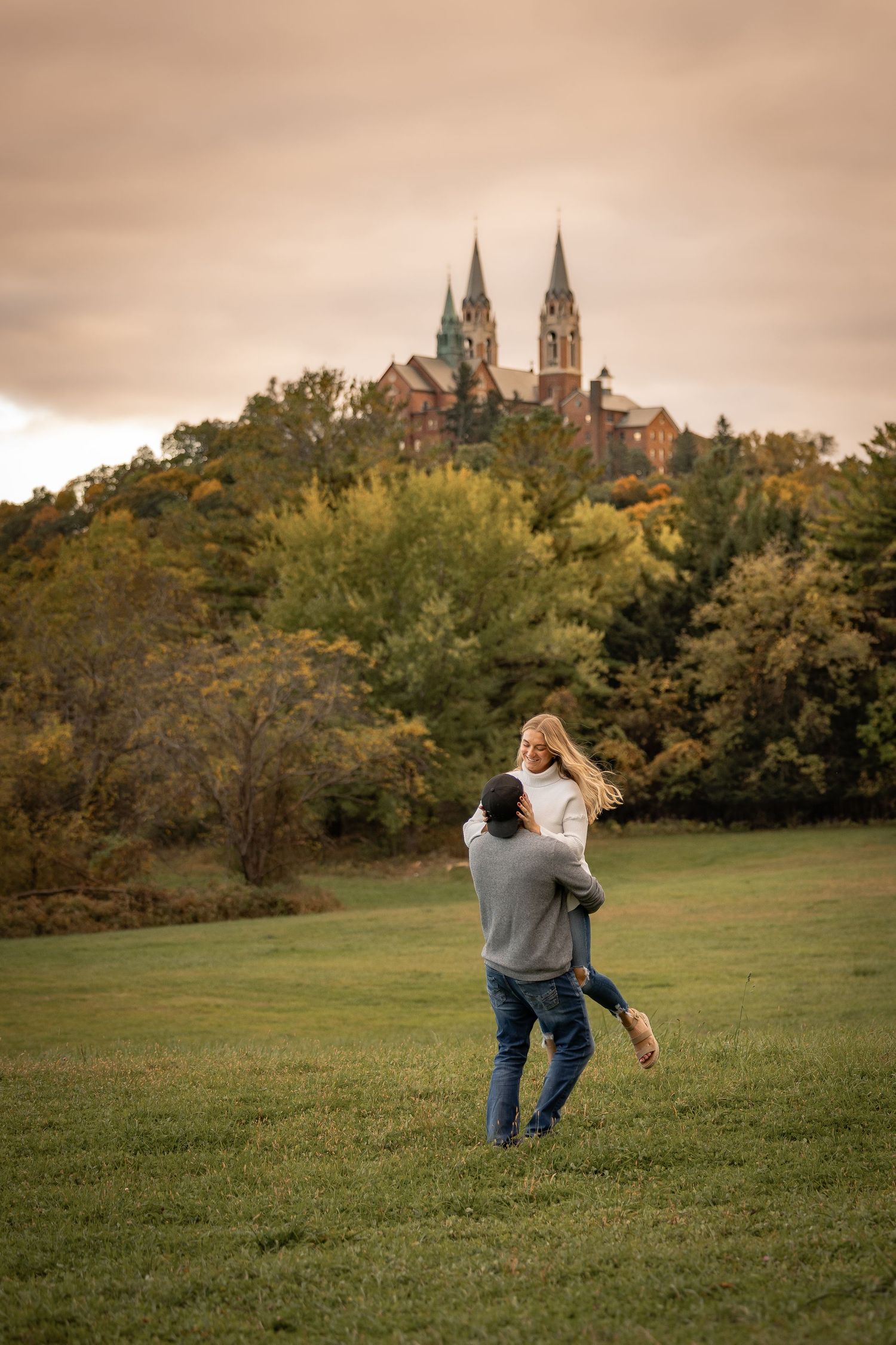 Couple spinning joyfully in green field with Holy Hill church landmark in autumn background.