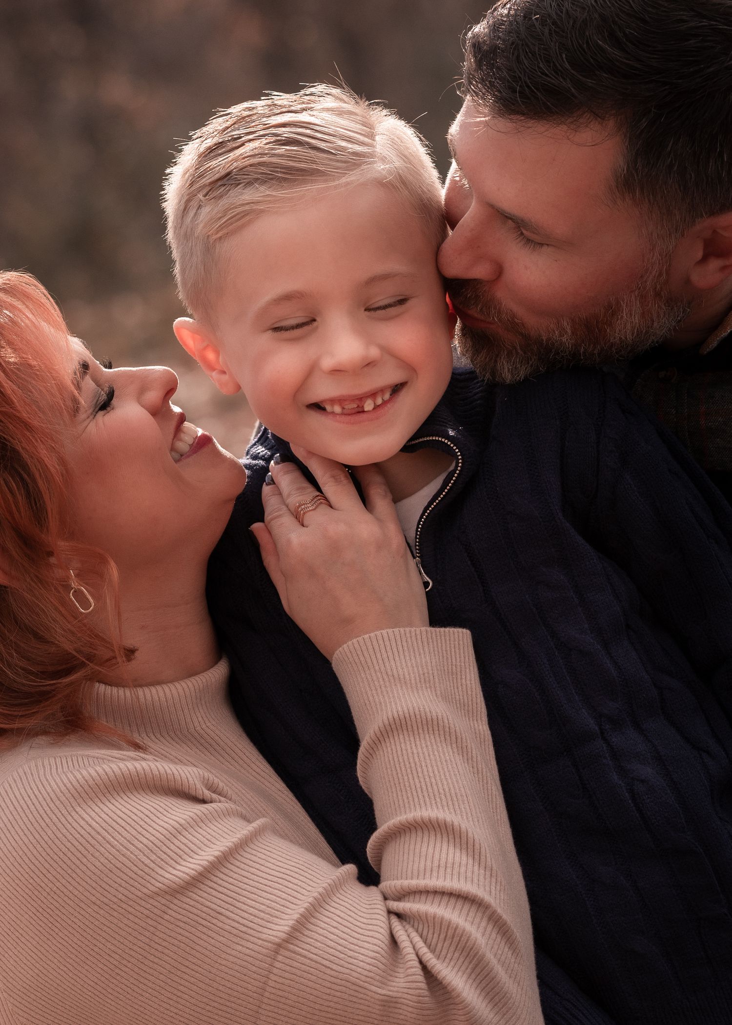 Parents lovingly kiss their smiling young blonde son in a warm outdoor family portrait.