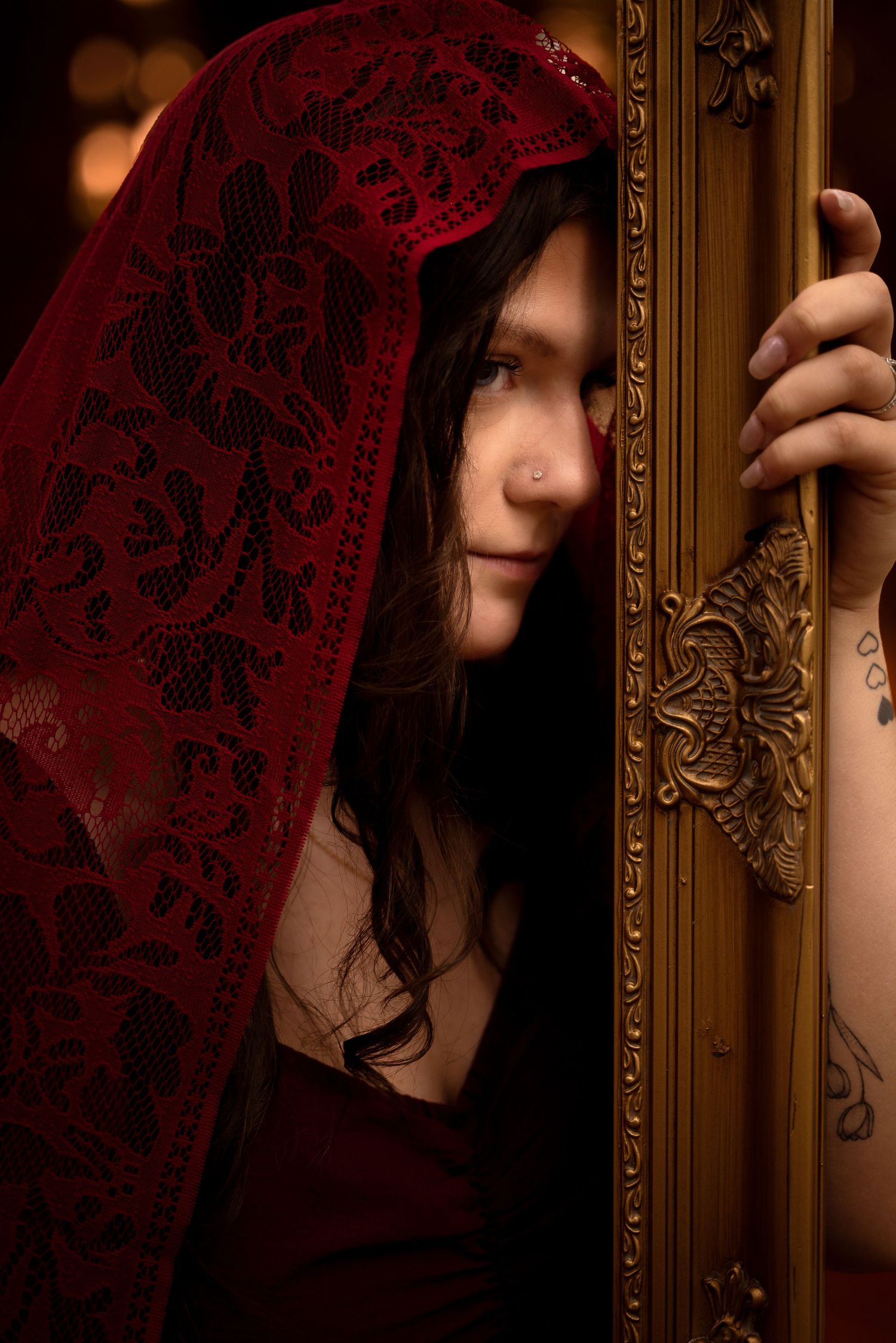 A mysterious woman in red lace veil peers around an ornate golden mirror frame in dramatic moody lighting.