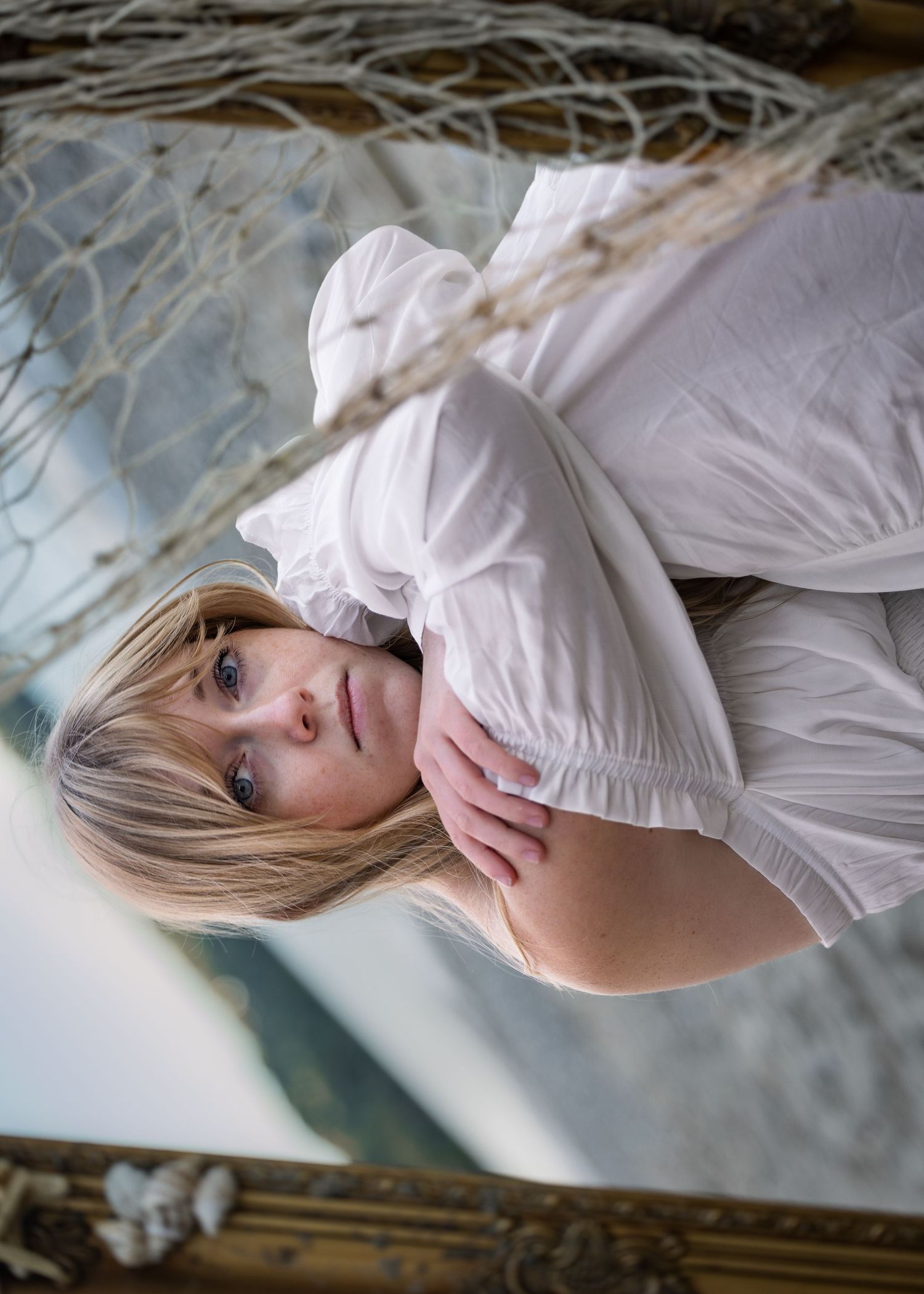 Blonde woman in white dress lying in a hammock near the beach, looking at camera.