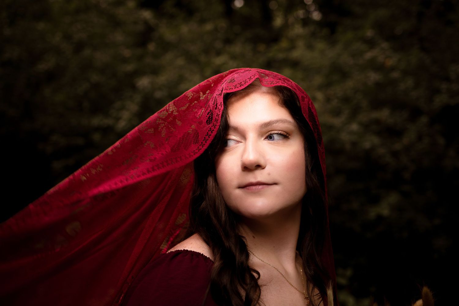 Young woman in red velvet hooded cape gazes upward outdoors, surrounded by lush green foliage.
