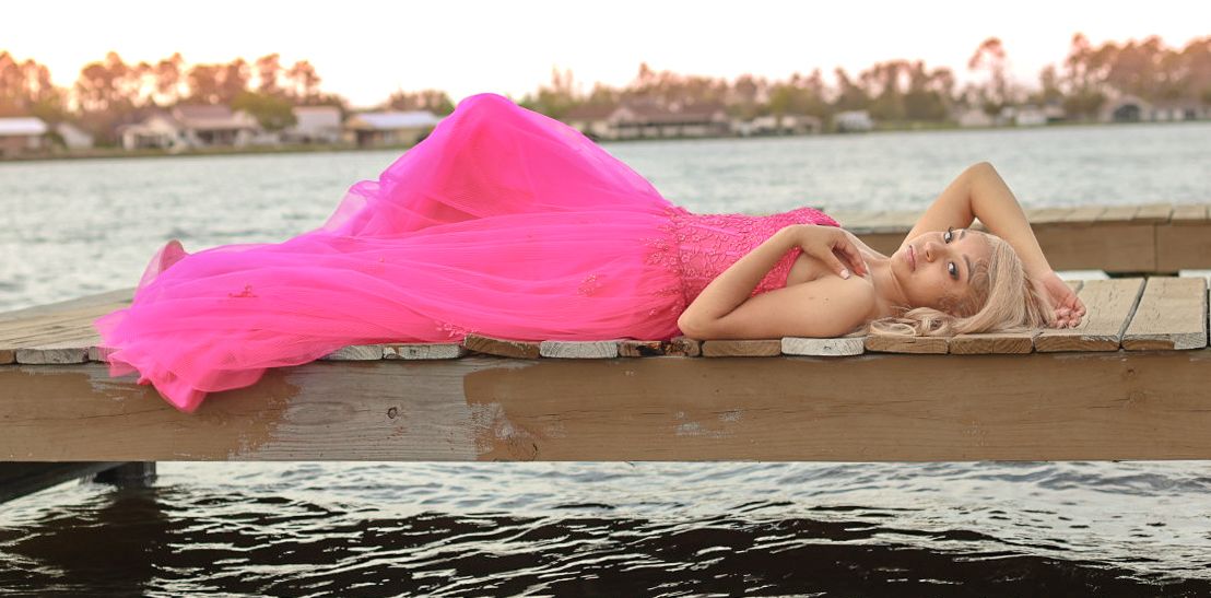 A flowing hot pink dress billows dramatically on a wooden dock at sunset over calm waters.