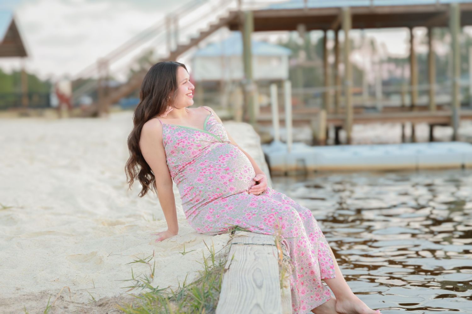 A woman in a pink floral dress poses gracefully by a lake during a golden hour photoshoot.