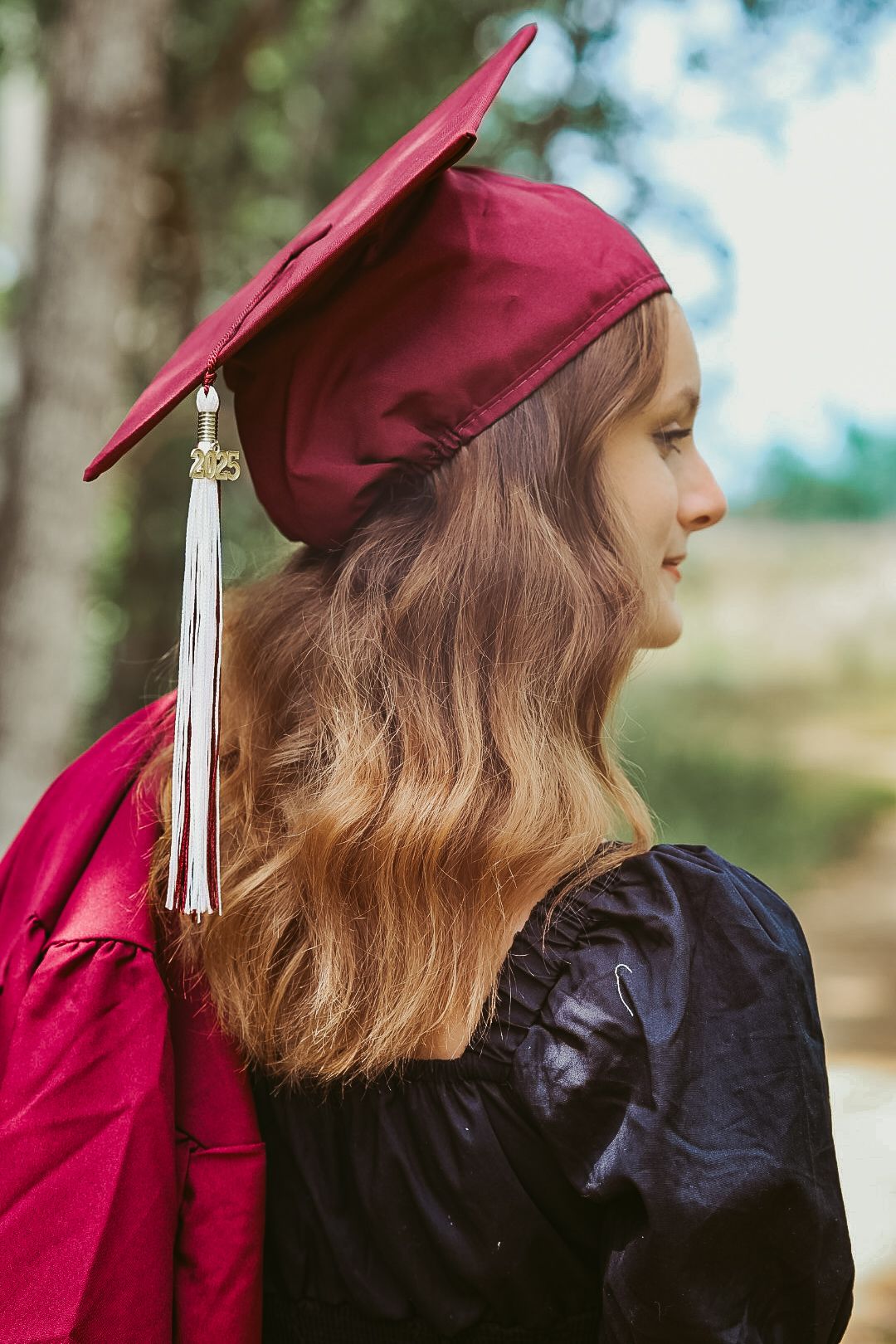 Young woman in maroon graduation cap and gown poses outdoors with long wavy hair and white tassel.