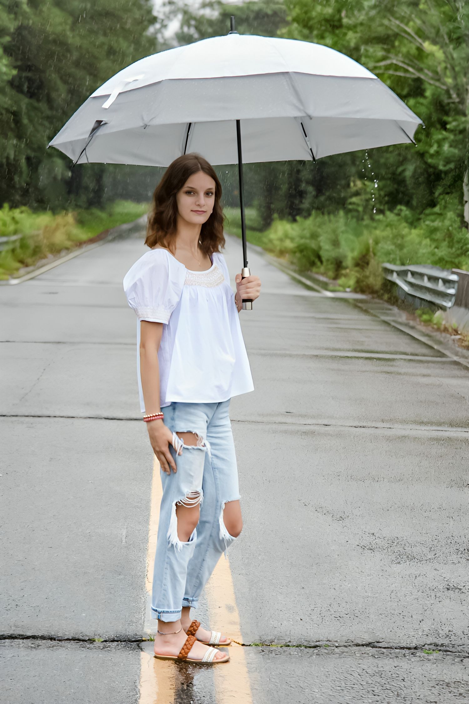 Young woman in white blouse and ripped jeans holding a white umbrella standing on a wet road in the rain.