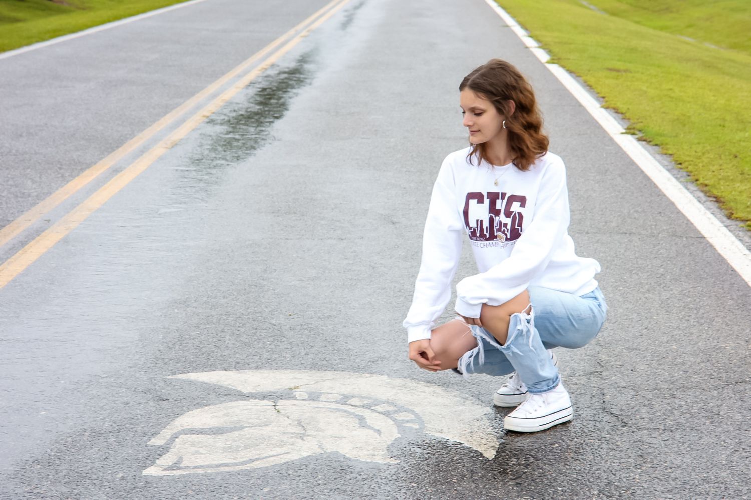 Teen girl in white CHS sweatshirt and jeans crouching on road examining chalk drawing on pavement.