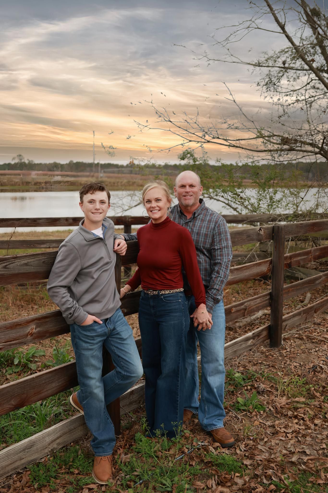 Family poses together on wooden fence at sunset by lake with autumn foliage in background.